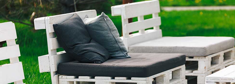 Sedie a sdraio da esterno realizzate con pallet di legno bianco impilati, con cuscini e guanciali grigio scuro su un prato verde.