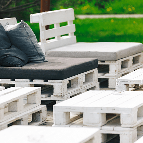 Seduta da esterno realizzata con pallet di legno dipinti di bianco, con cuscini grigio scuro su uno sfondo di prato.