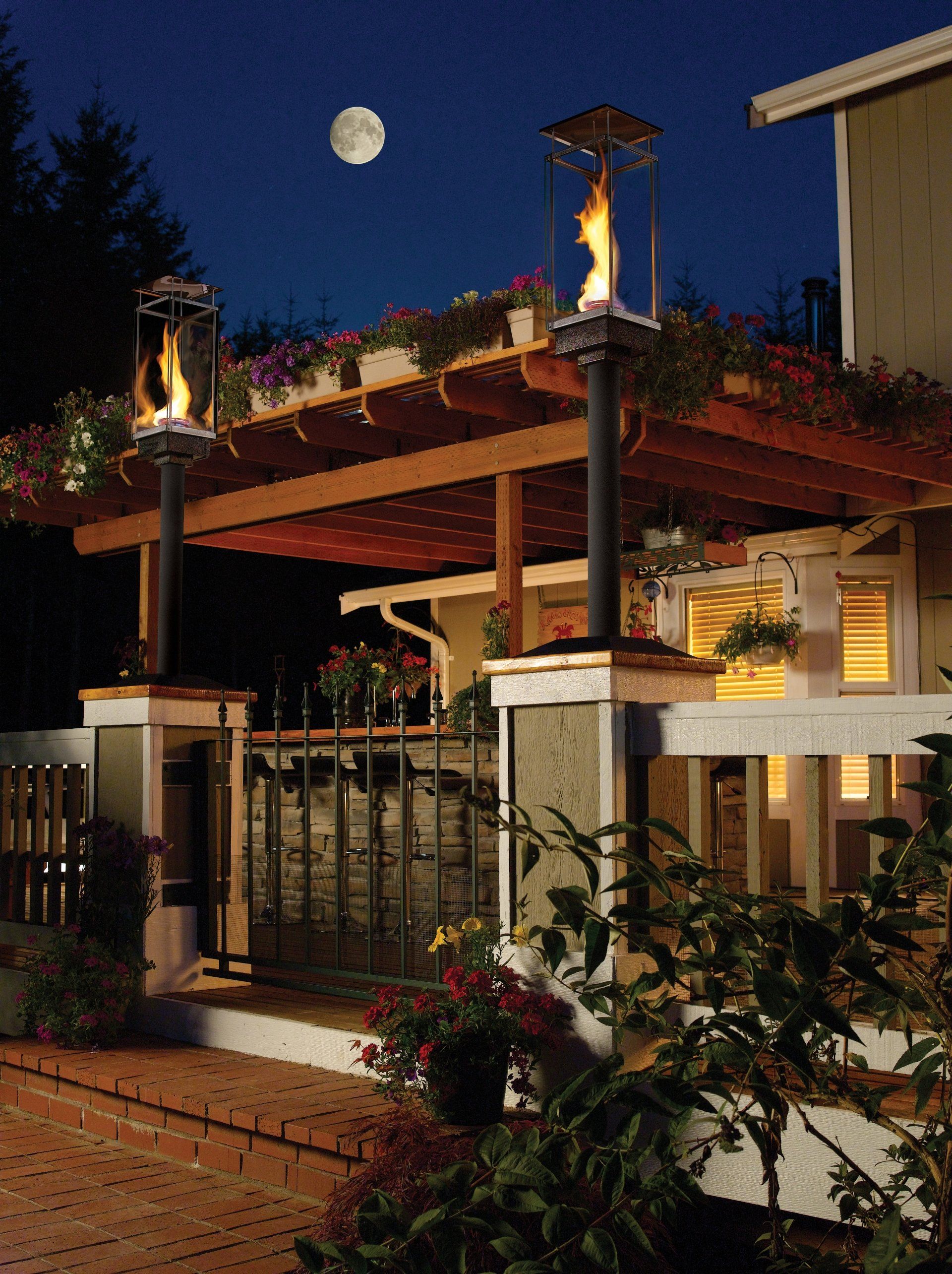 Two outdoor gas lamps with flames, pergola with flowers, under a moonlit sky.