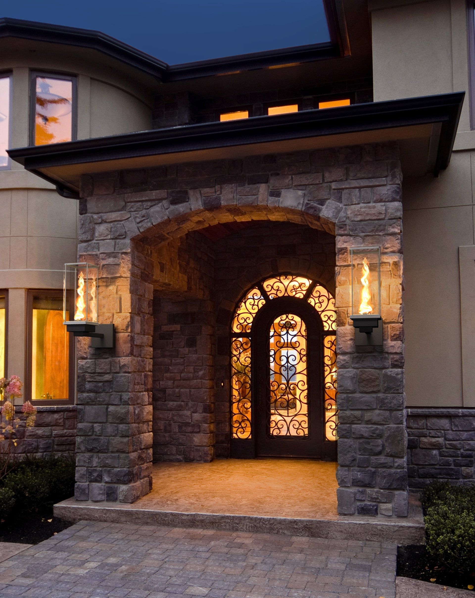 Stone arched entry to a house with a decorative glass door, lit by sconces with flames.
