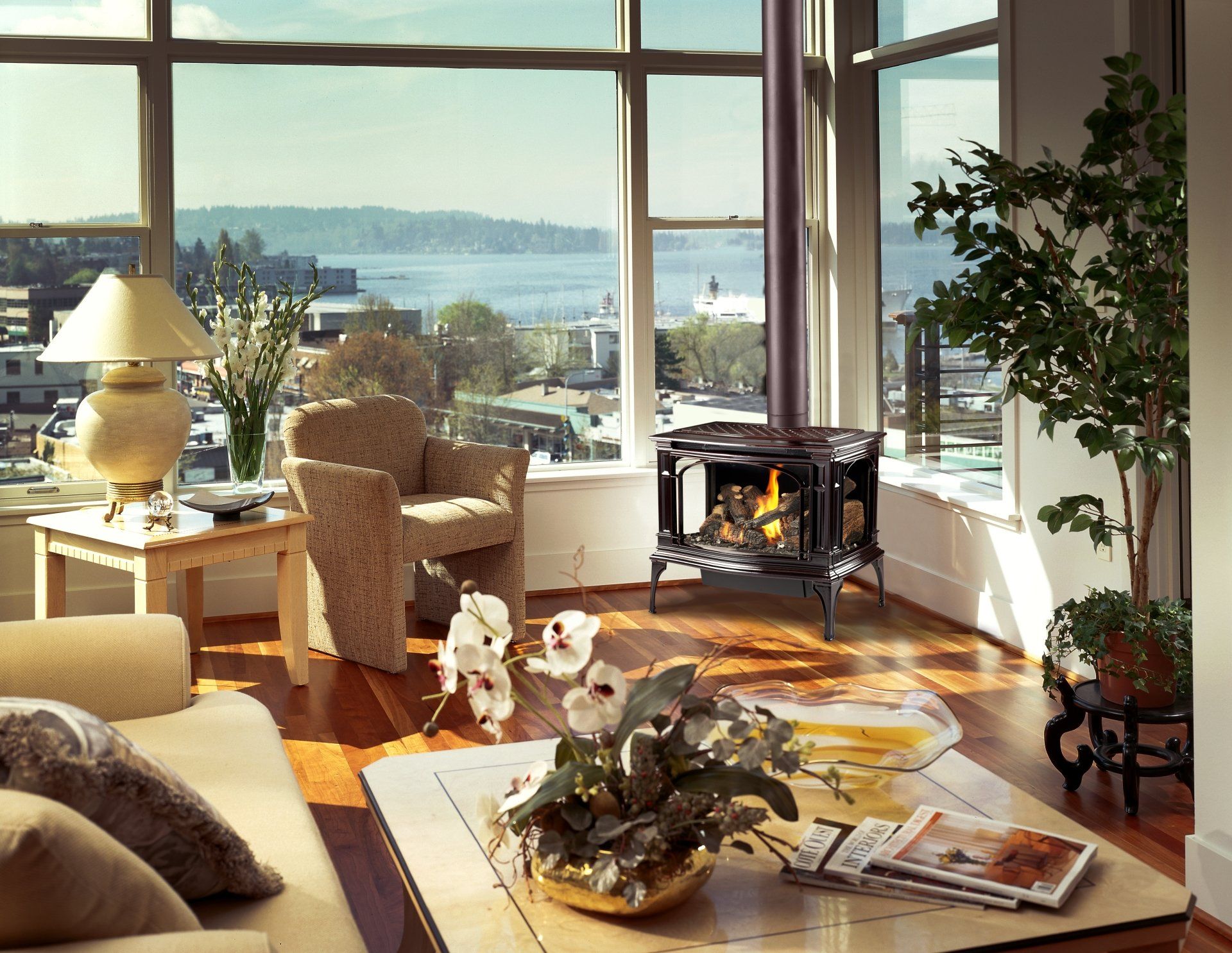 Cozy living room with wood-burning stove, large windows, city view, armchair, sofa, and plants.