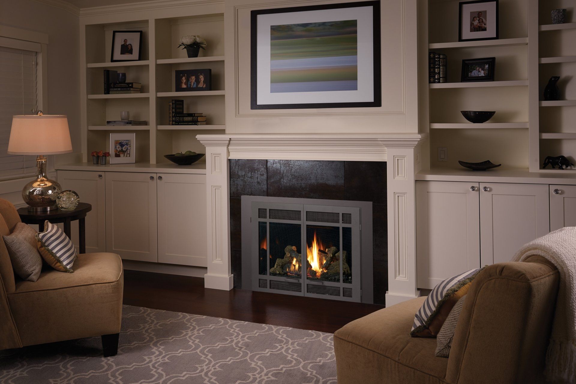 Cozy living room with fireplace, built-in shelving, two armchairs, and a lit lamp. Warm tones and soft lighting.