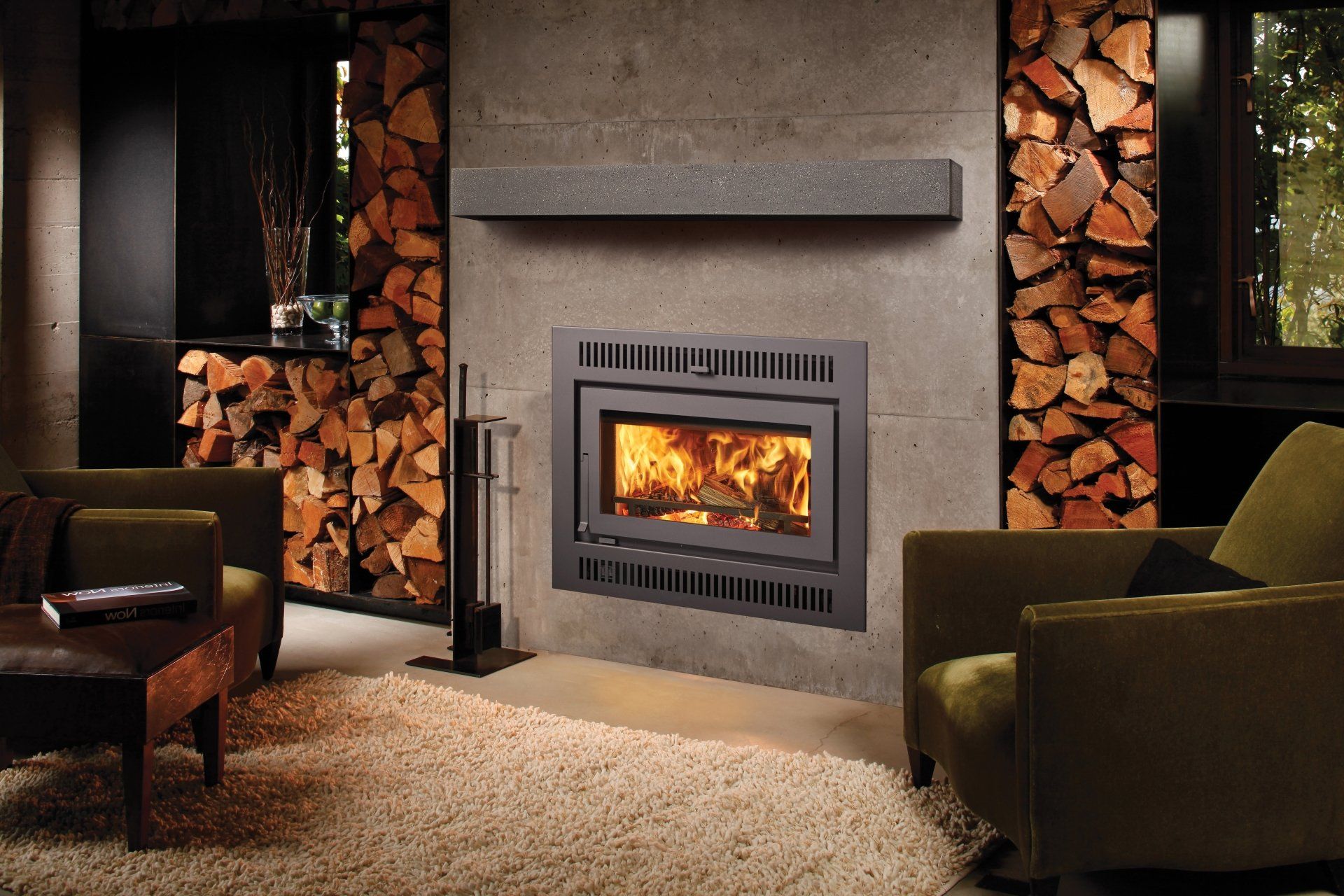 Cozy living room with fireplace, firewood stacks, and two olive green armchairs on a beige carpet.