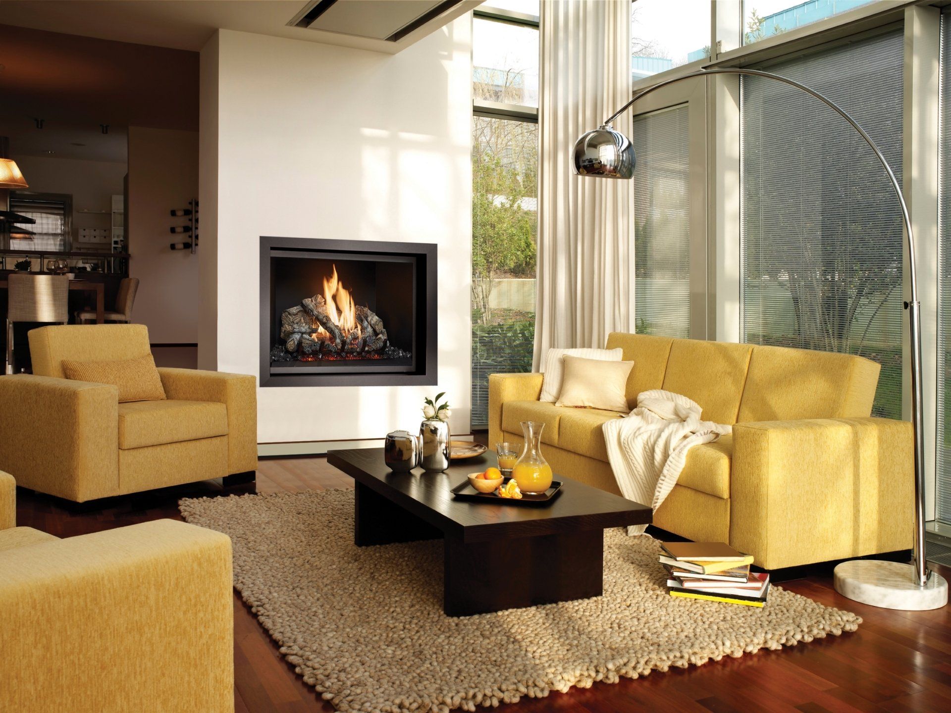 Cozy living room with fireplace, yellow sofa/armchairs, coffee table, rug, hardwood floors, and large windows.
