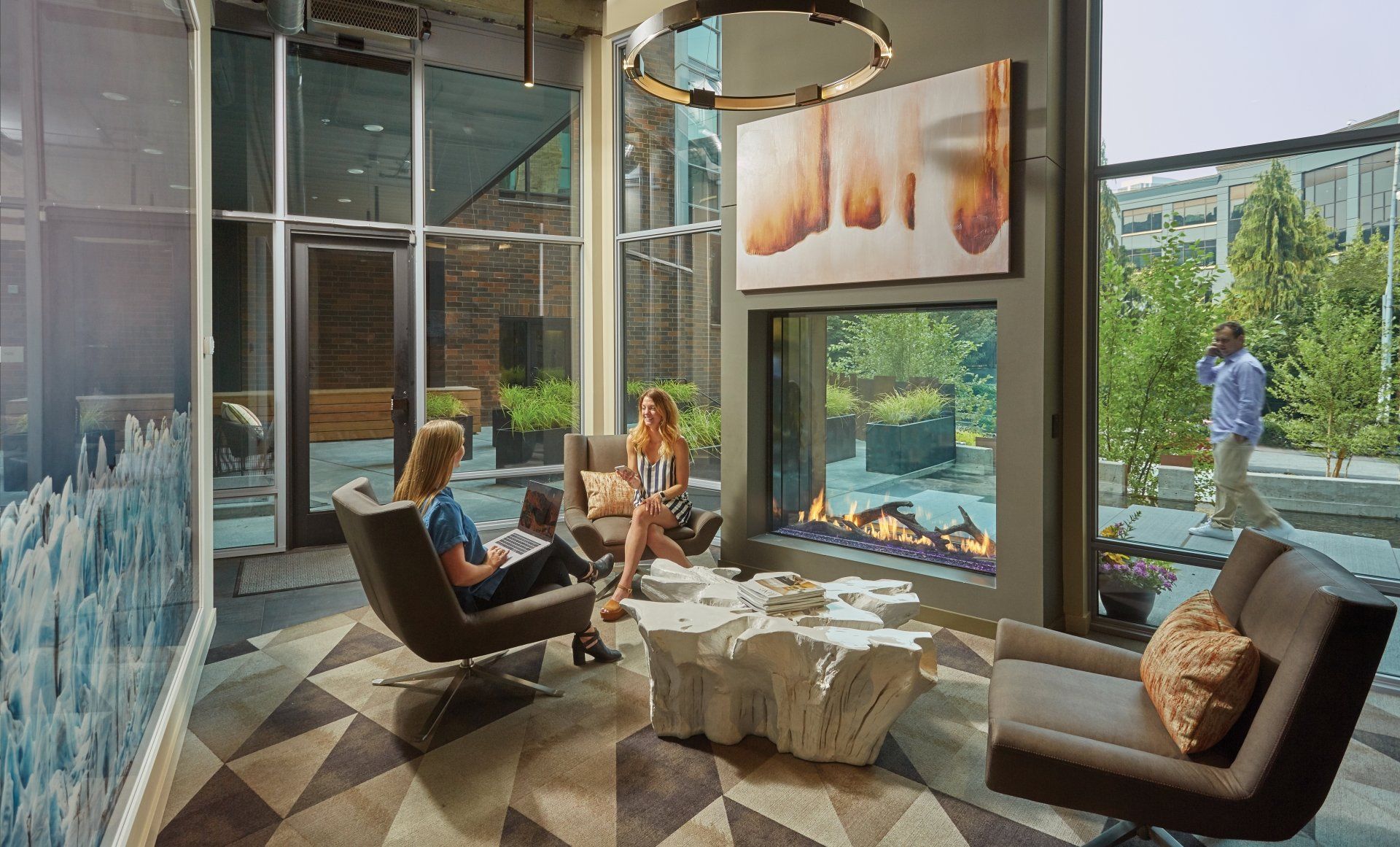 Two people in lounge chairs by fireplace, looking at phones. Modern interior with art and outdoor view.