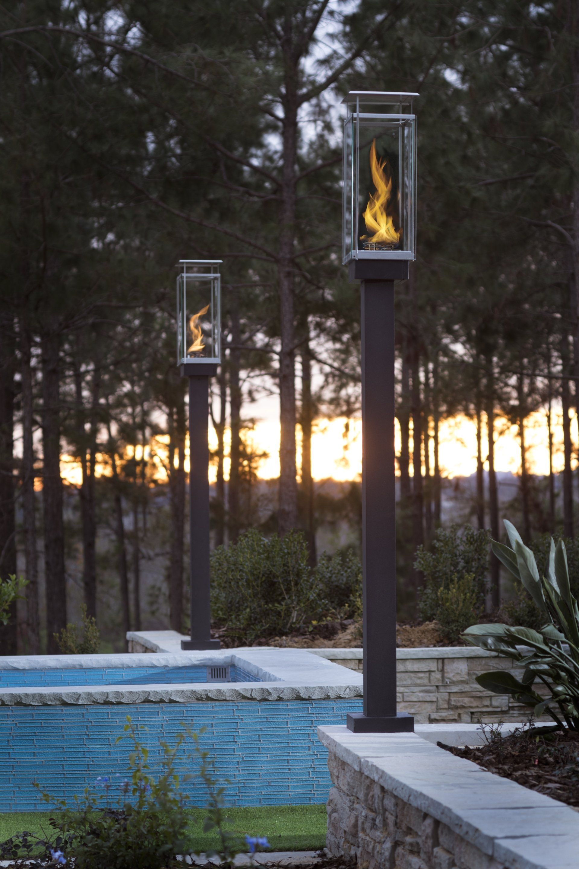 Two outdoor gas fire pits illuminate a pool area in front of a forest at sunset.