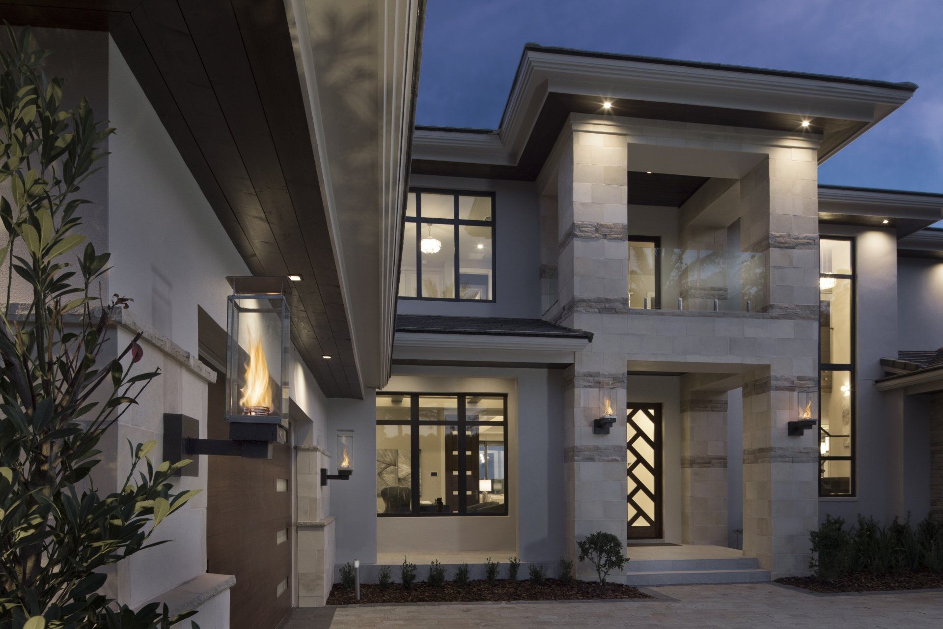 Modern two-story house with stone facade, glass windows, and outdoor fire feature, lit up at dusk.