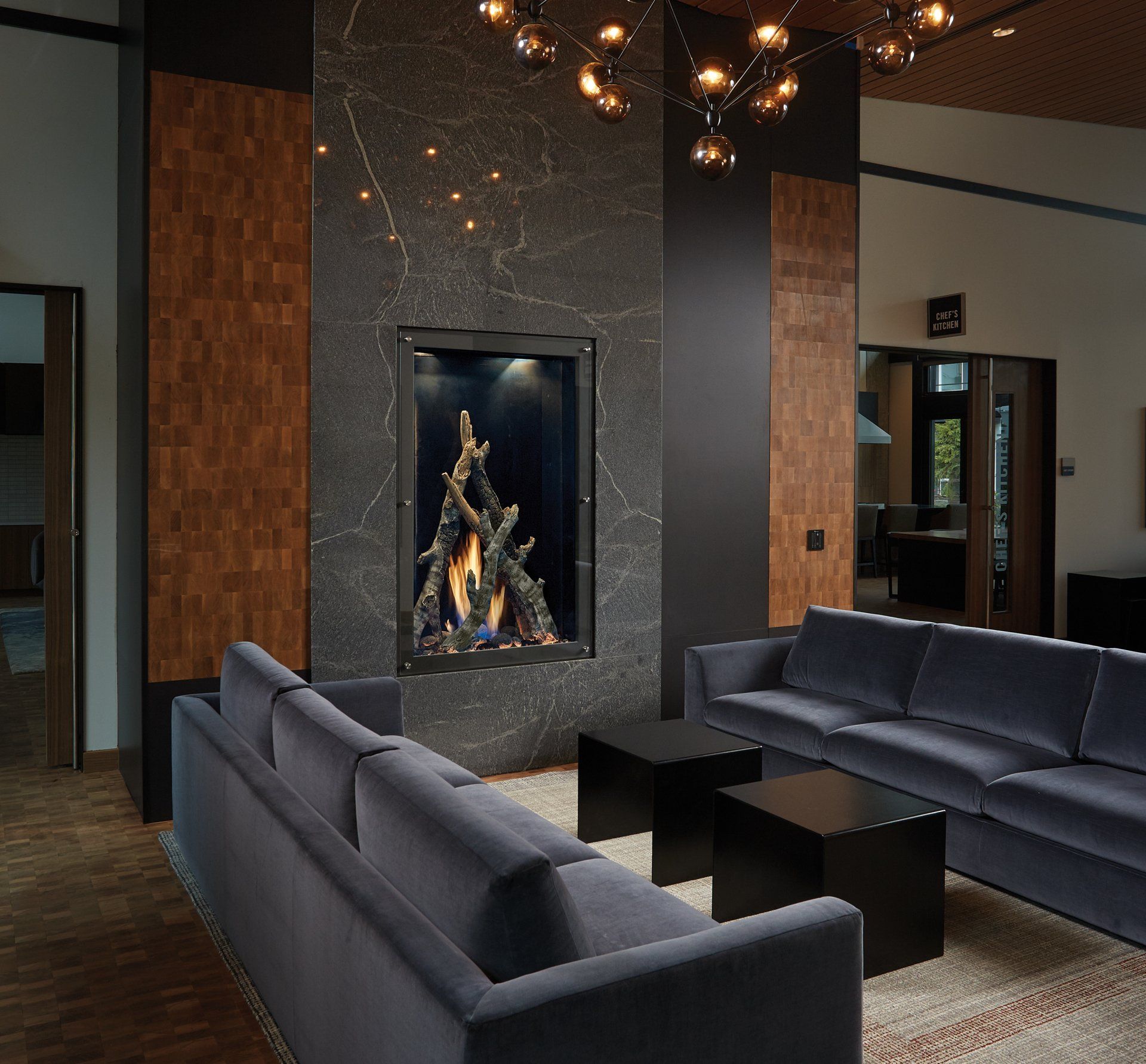 Modern living room with a fireplace, dark furniture, and a decorative light fixture.