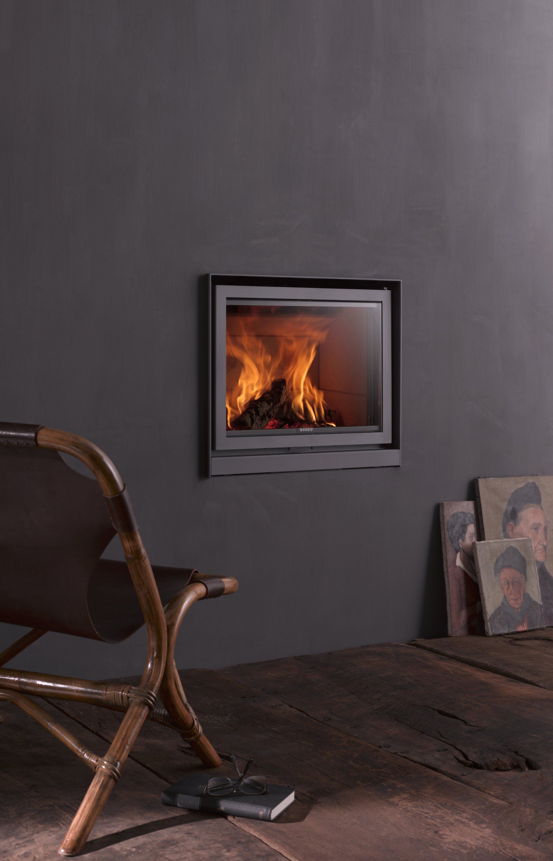 Cozy fireplace inset in dark gray wall with chair and wooden floor. Burning flames inside the fireplace.
