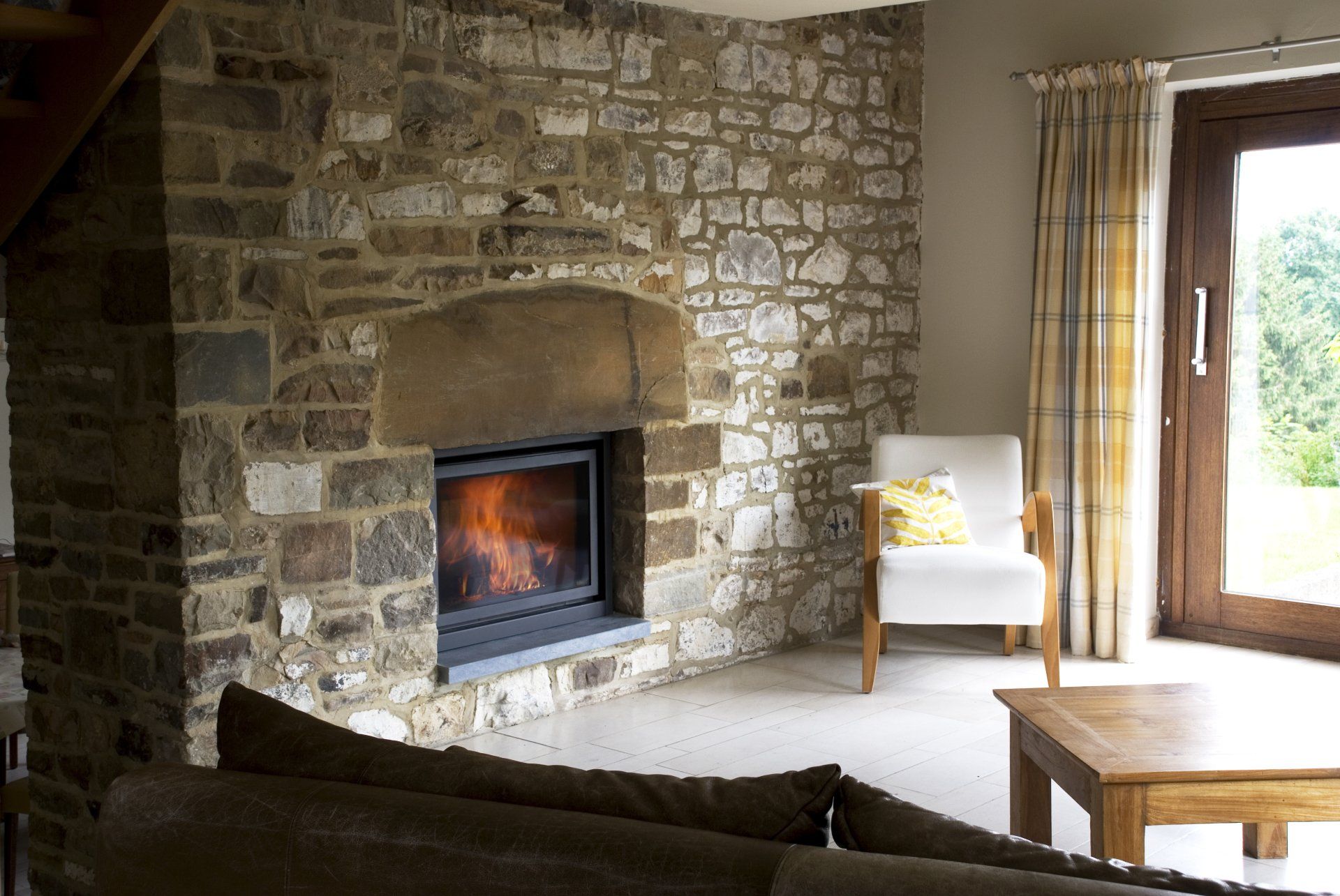Stone fireplace with a fire, living room with a chair, and wooden door to a green view.