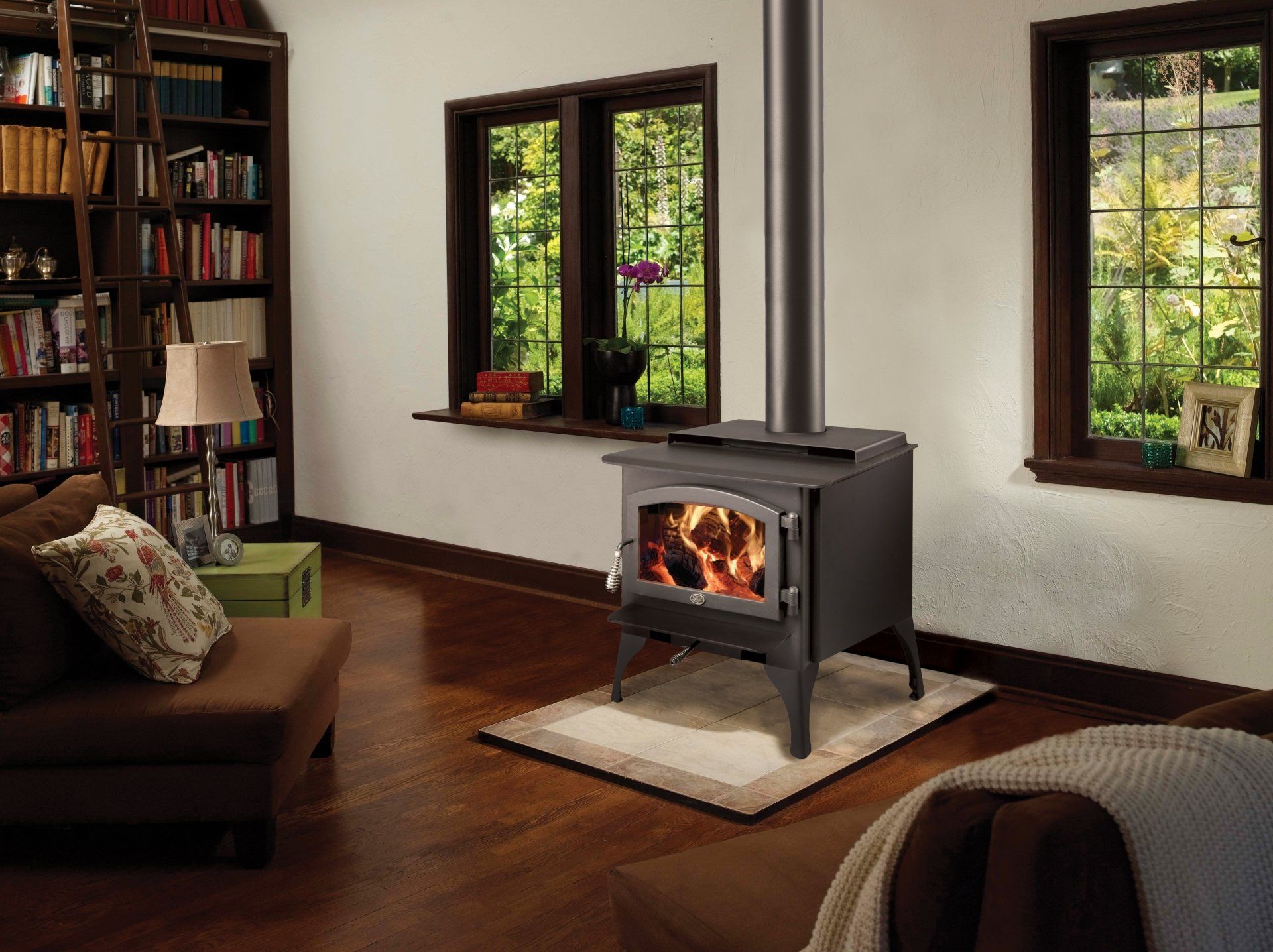 Cozy living room with a lit wood stove. Windows frame a lush outdoor view, a bookshelf is visible to the left.