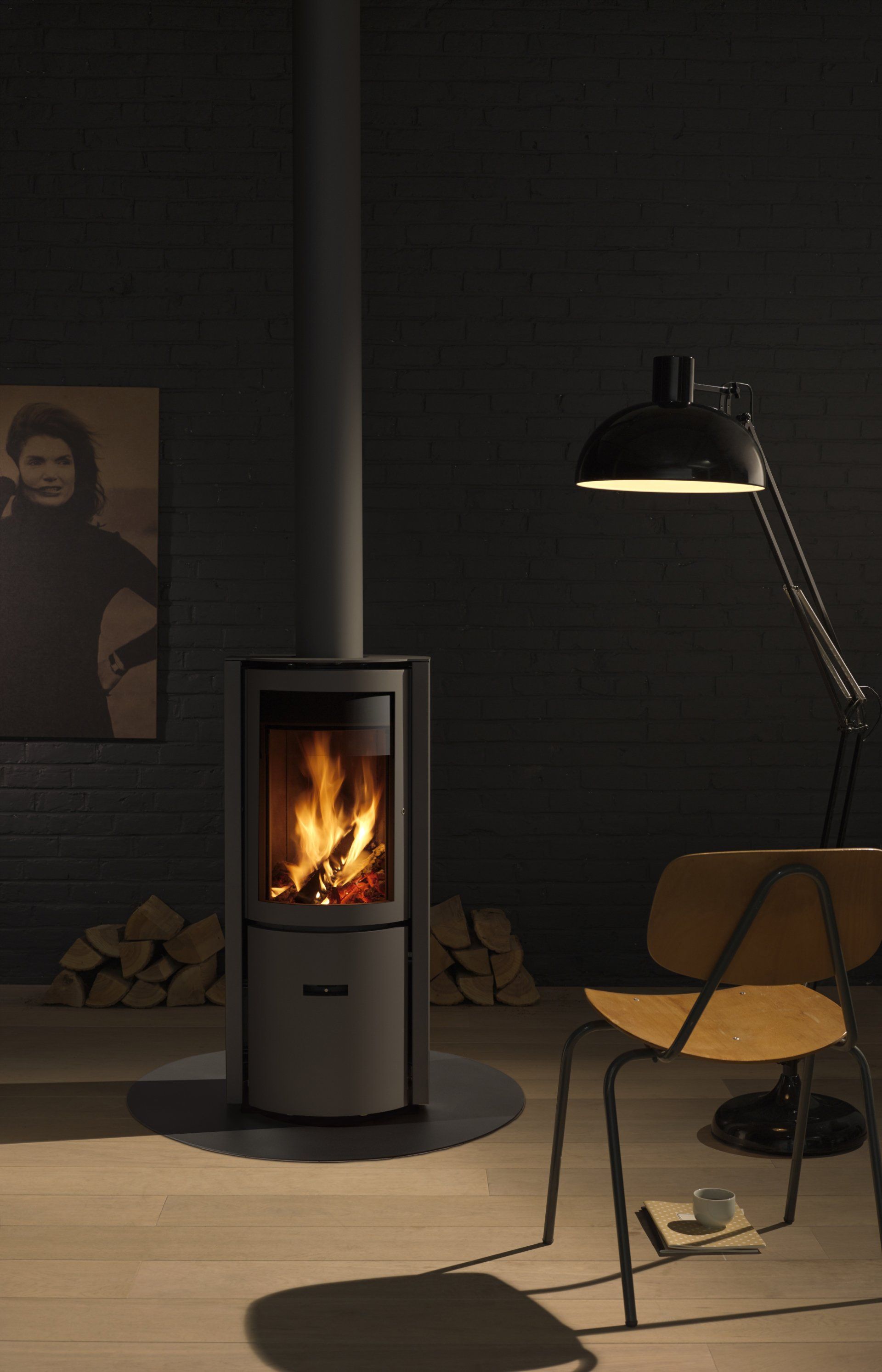 A lit fireplace in a gray stove with a dark room, chair, and a picture on the wall.