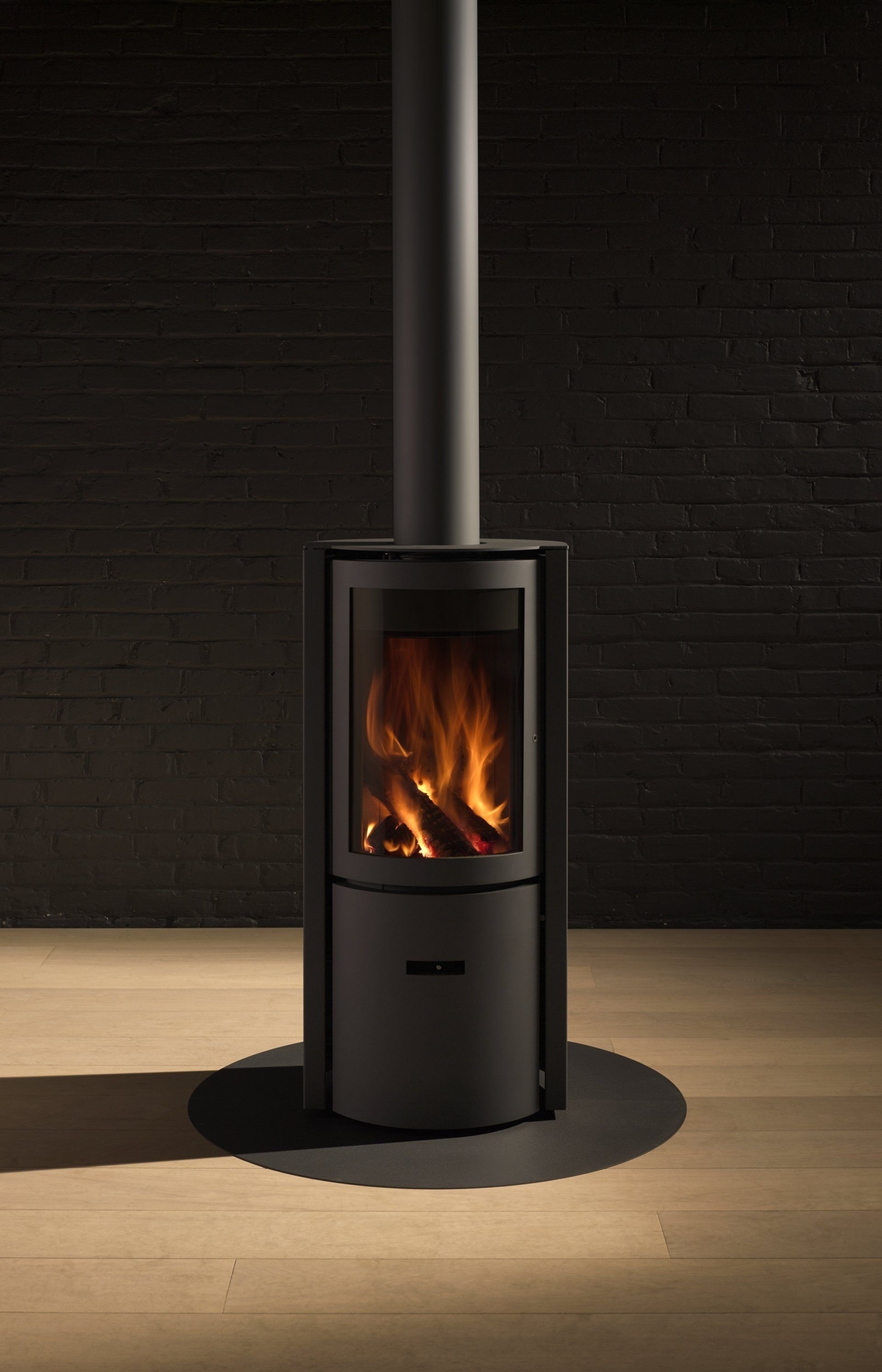 Black wood-burning stove with flames, black chimney pipe, on wooden floor against a dark brick wall.