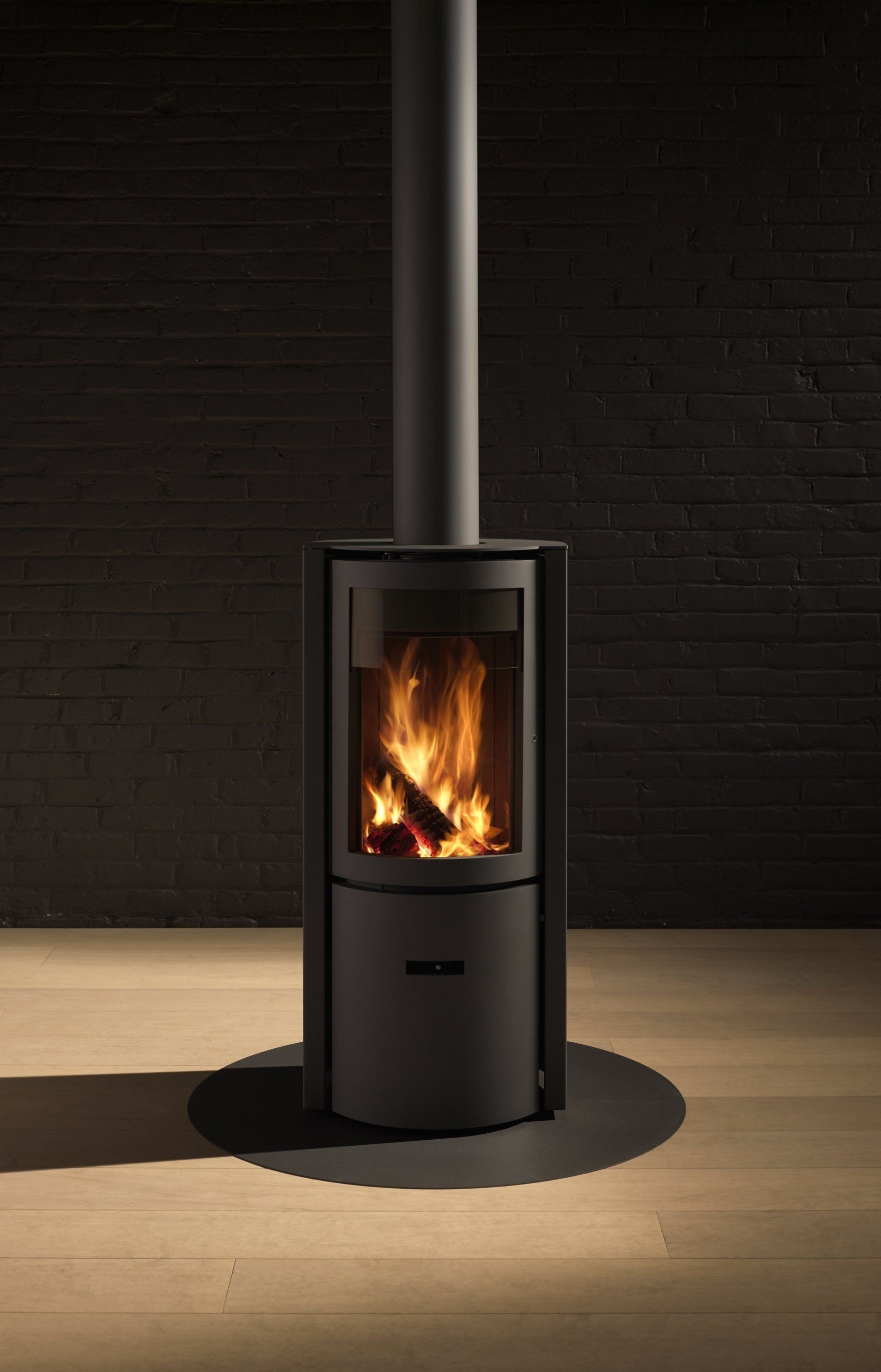 Black wood-burning stove with flames, black flue, on wooden floor against a dark brick wall.