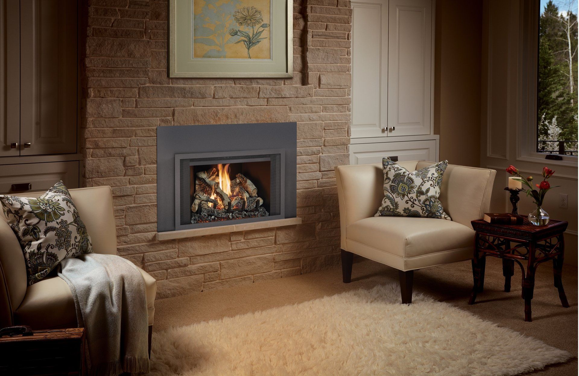 Fireplace in stone wall, flanked by cream armchairs, rug, and cabinets. Artwork hangs above the fireplace.