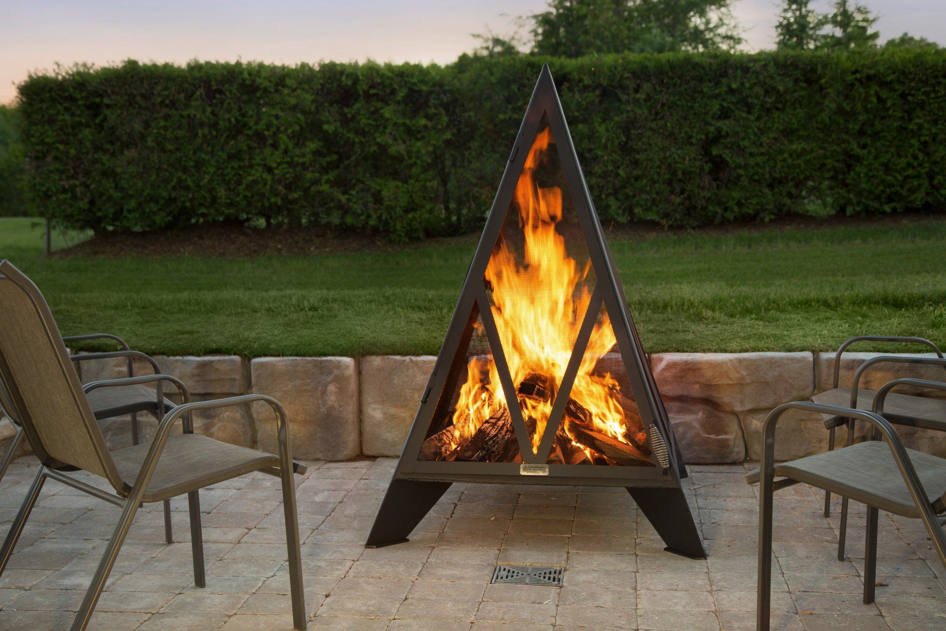 Triangular black fire pit with burning wood, surrounded by chairs on a patio, green hedge background.