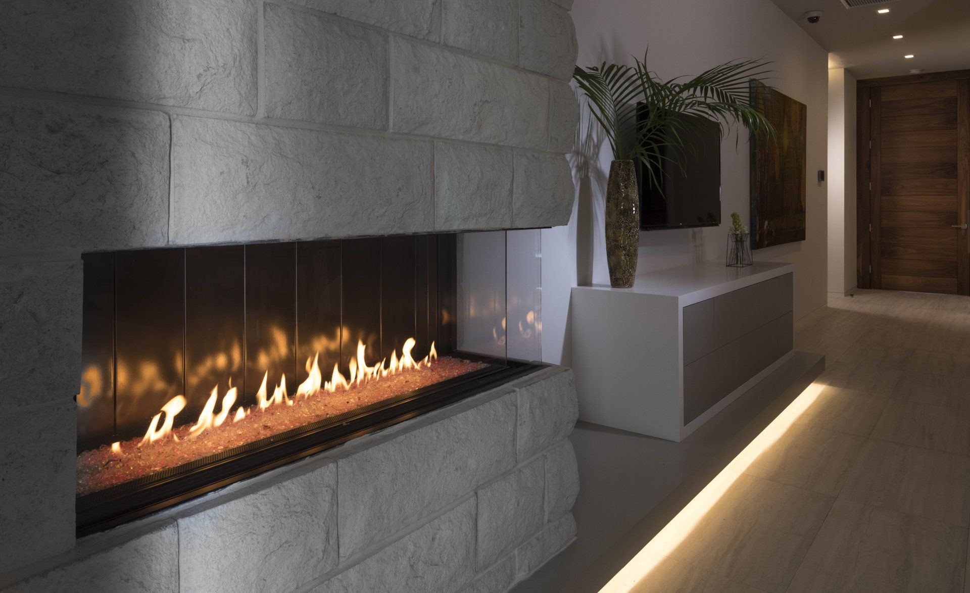 Modern fireplace with flames, built into a light gray stone wall. A TV and a plant are nearby.