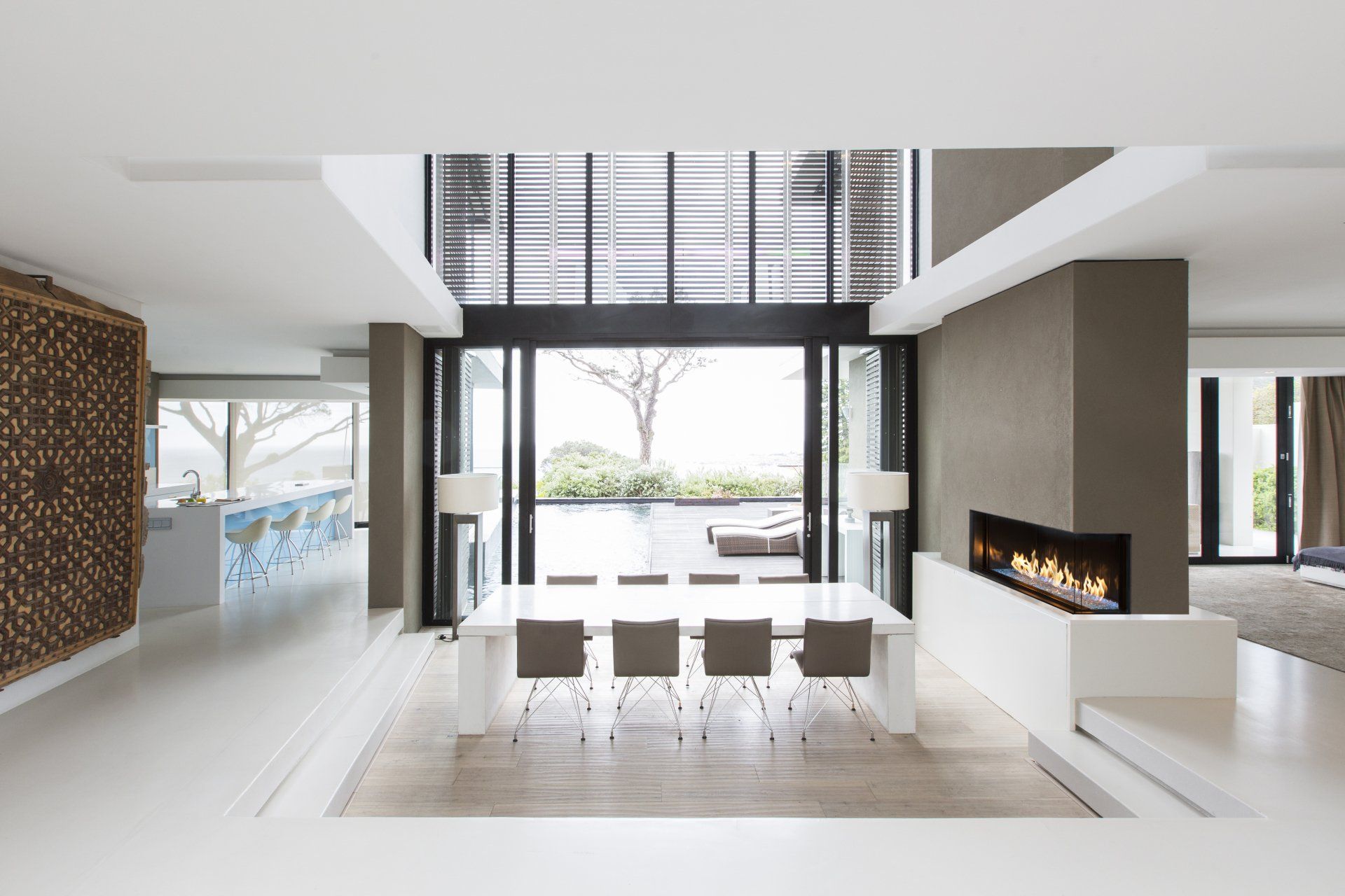 Modern dining room with white table, gray chairs, fireplace, and open glass doors.