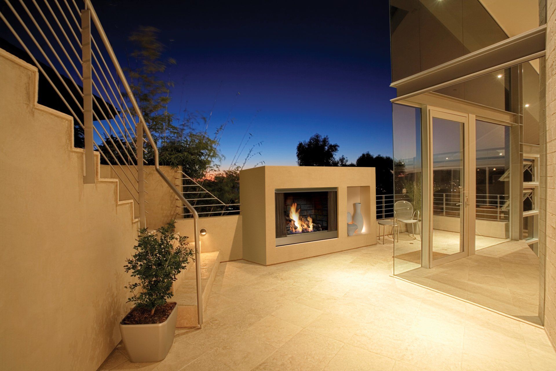 Patio with lit fireplace and stairs at dusk. Building with glass windows to the right.