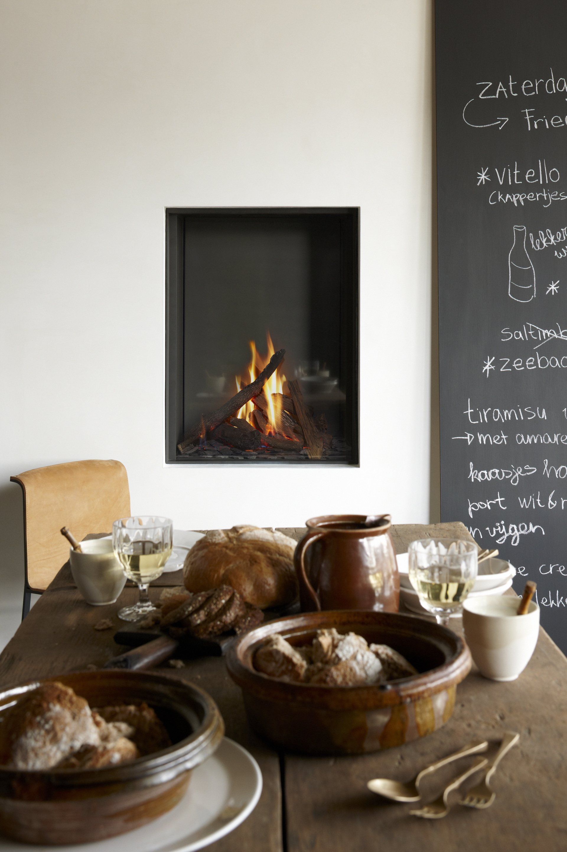 Rustic dining scene: Bread, stew, wine on a wooden table near a fireplace with flames. Chalkboard wall.