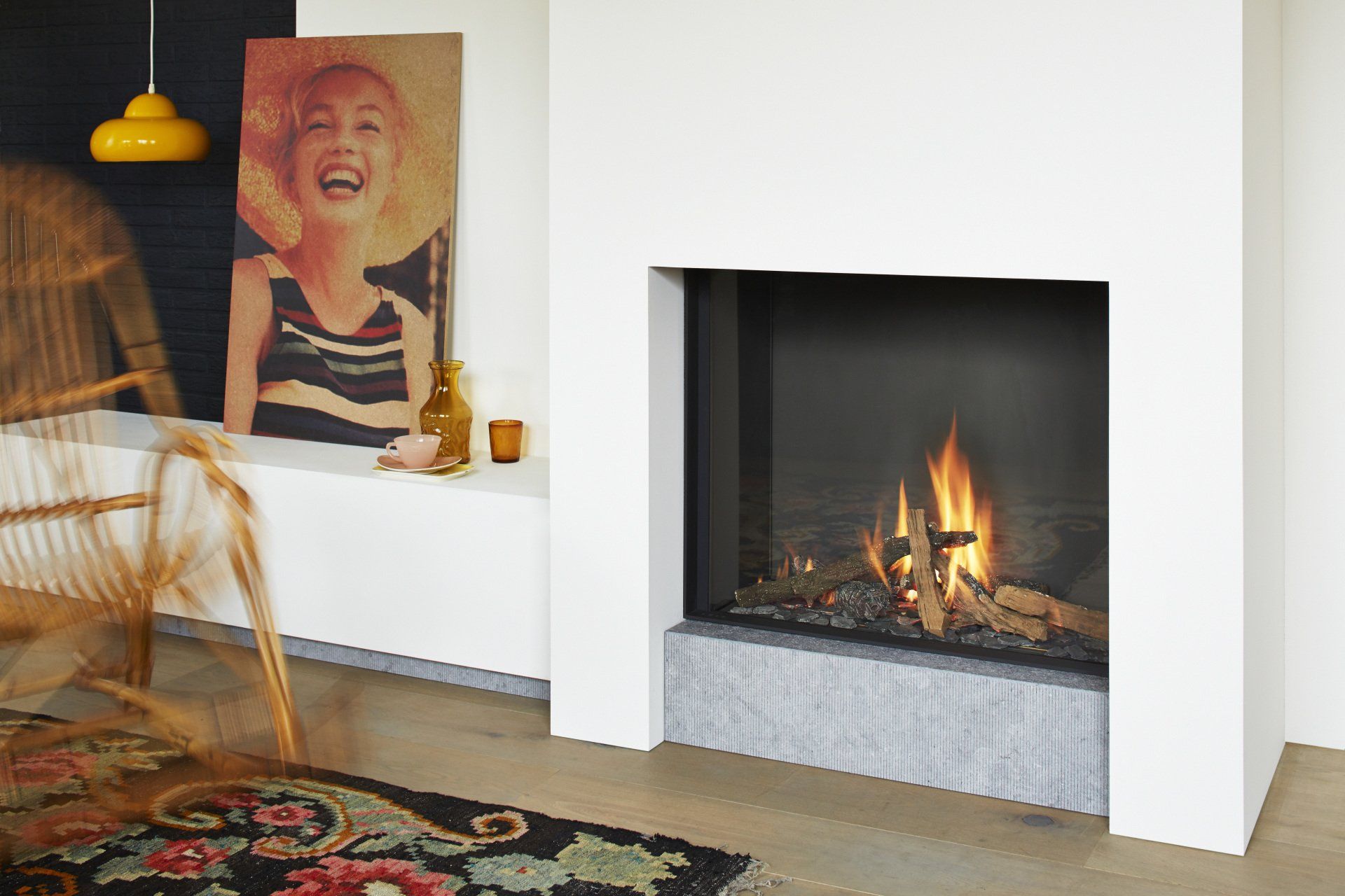 Modern fireplace with flames, gray stone base, white surround, artwork, and a yellow lamp.