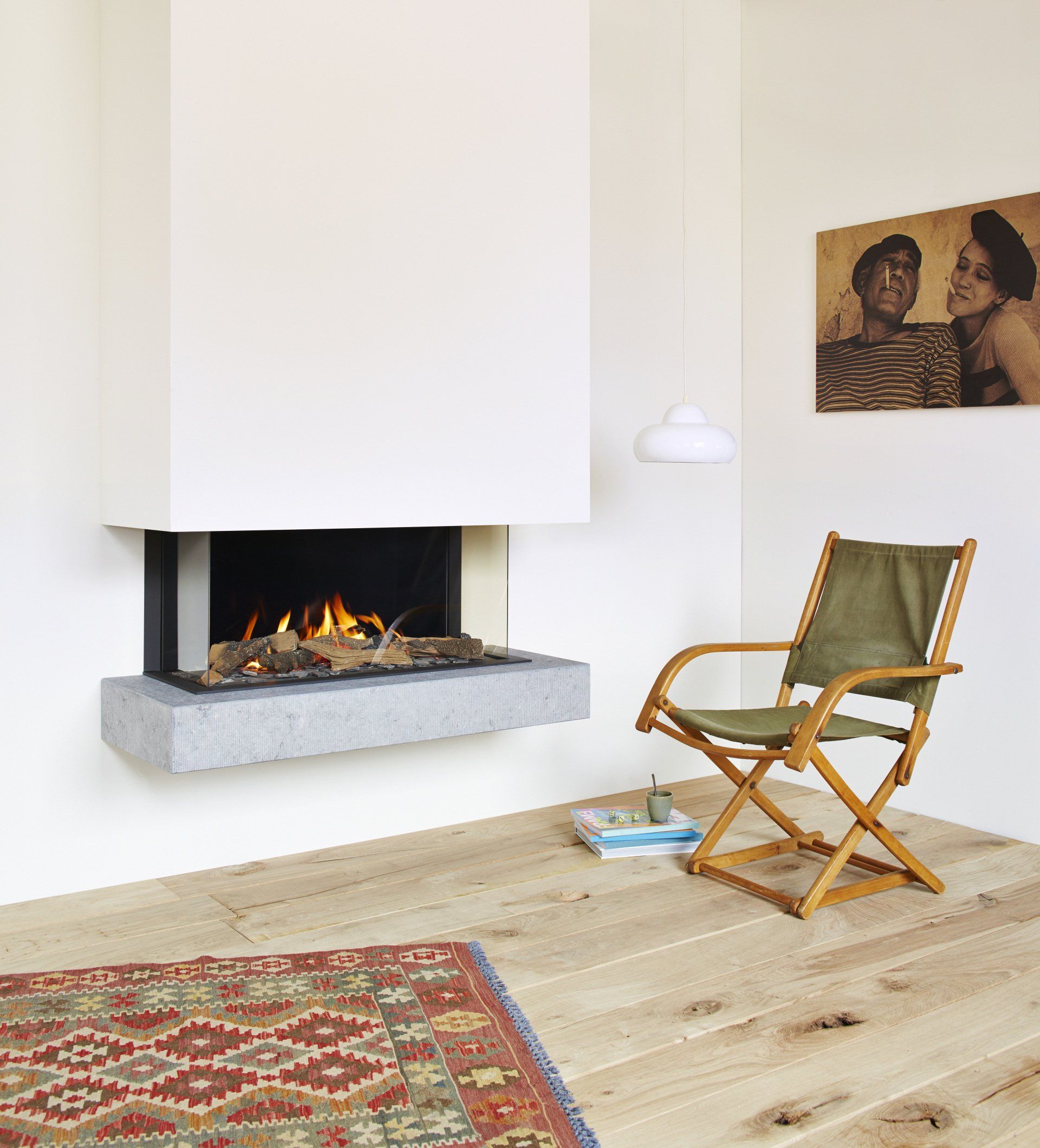 Fireplace with flames and wooden logs, a chair, rug, artwork, and light-colored wooden floors.