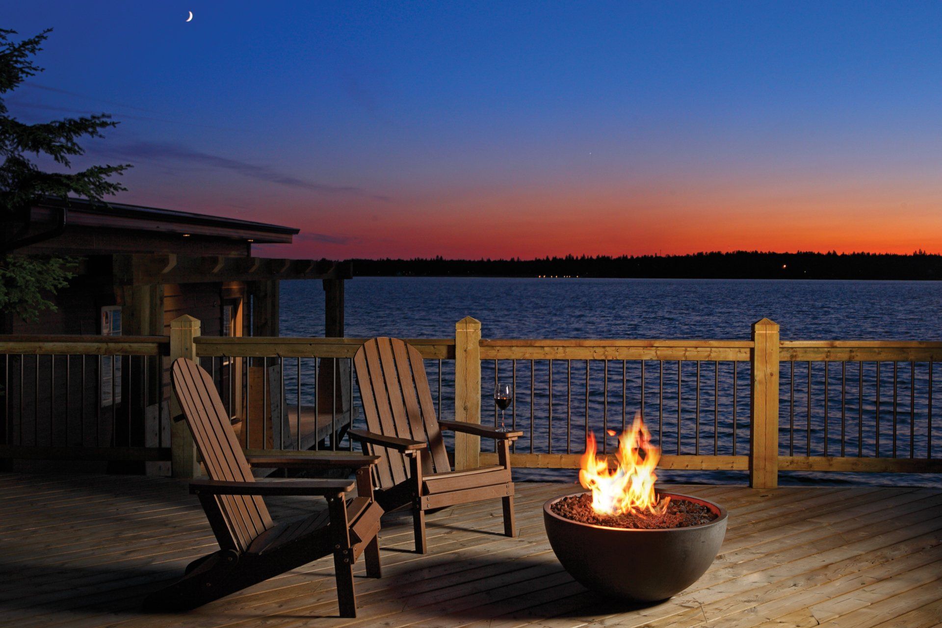 Two wooden chairs on a deck overlook a lake at sunset with a fire pit burning.