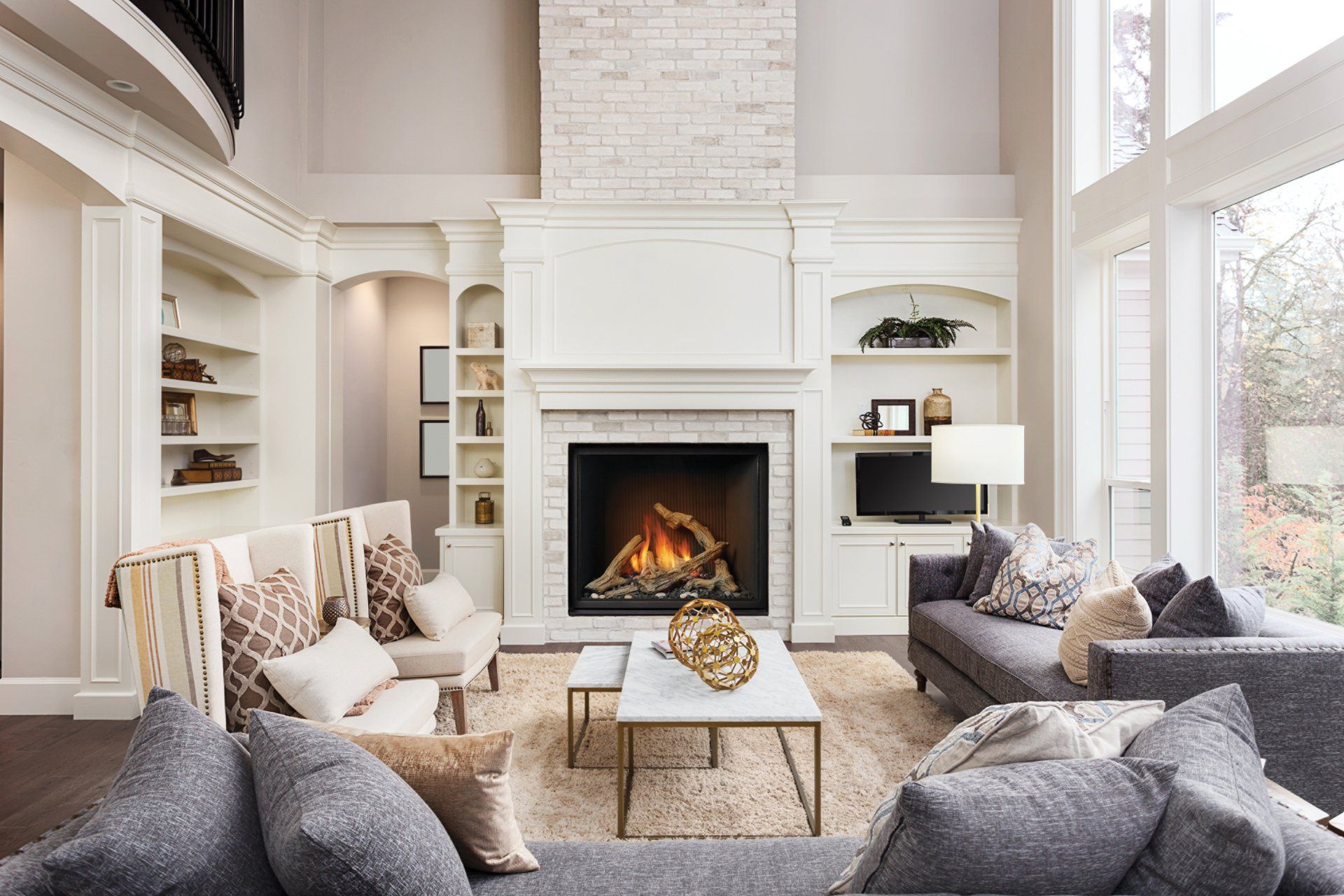 Living room with fireplace, built-in shelving, sofas, and large windows. Neutral color scheme, elegant setting.