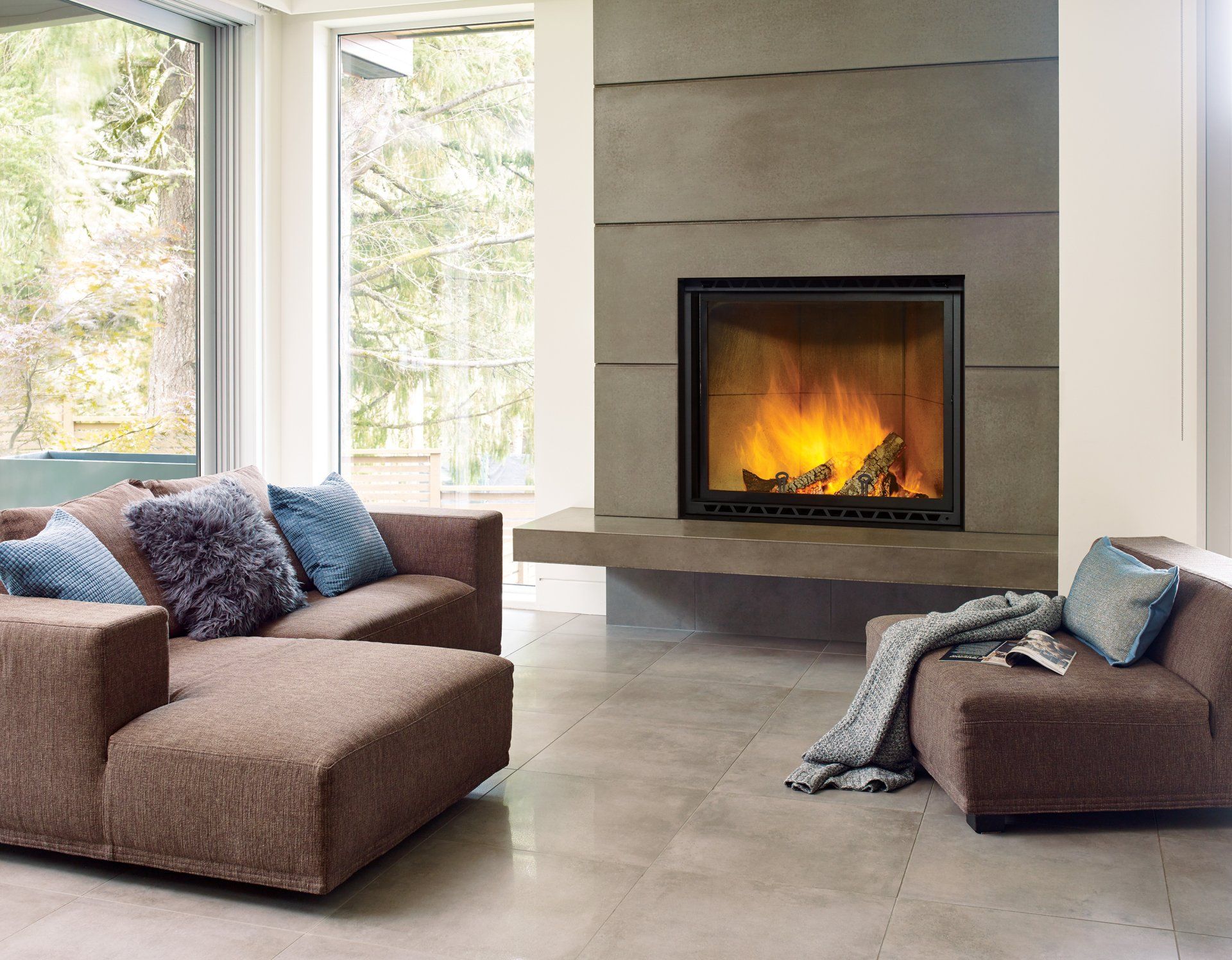 Living room with modern fireplace, brown couches, stone flooring, large windows, and lit fire.