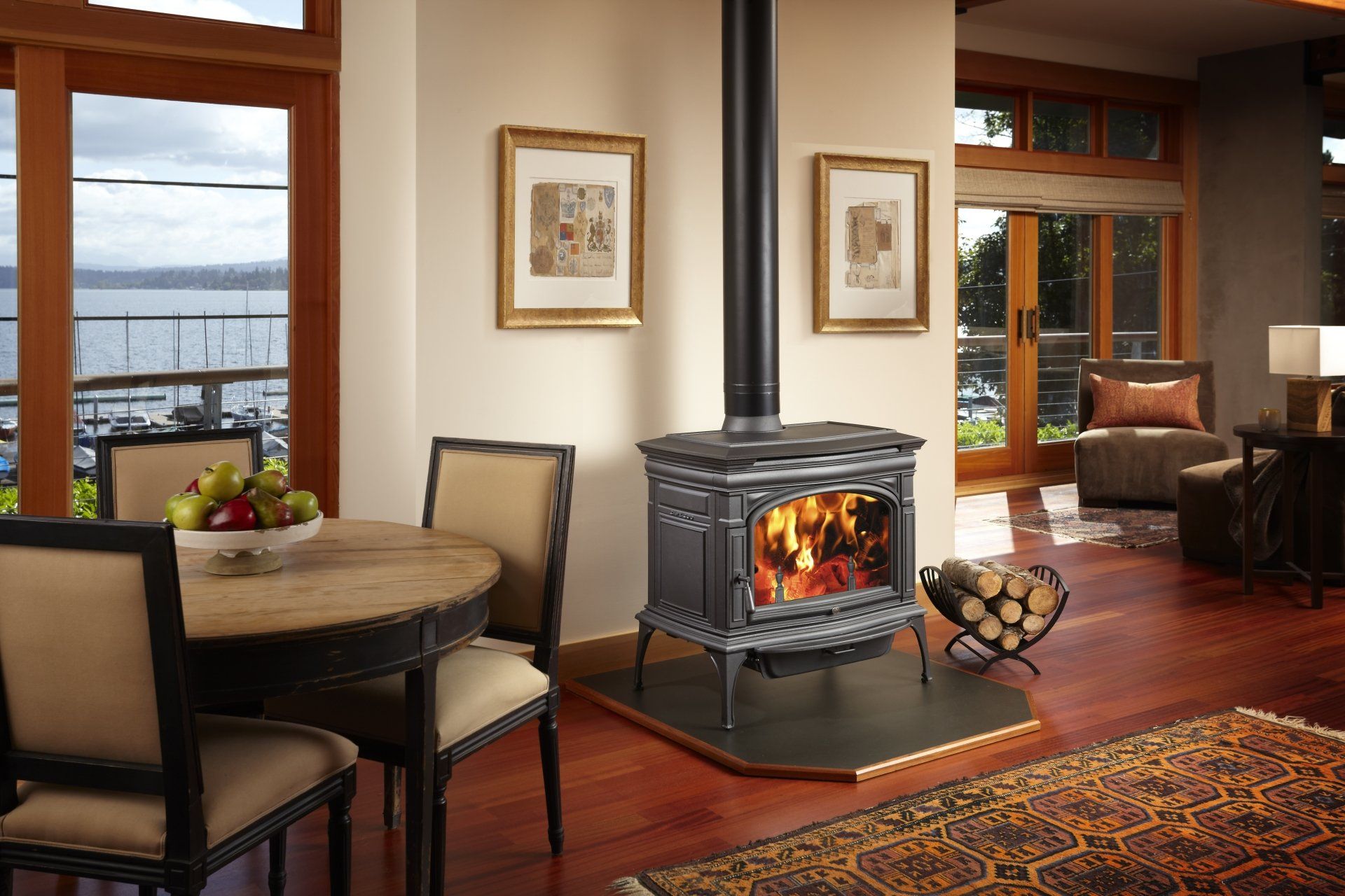 Cozy living room with a lit wood-burning stove; round table with fruit; windows overlooking water.