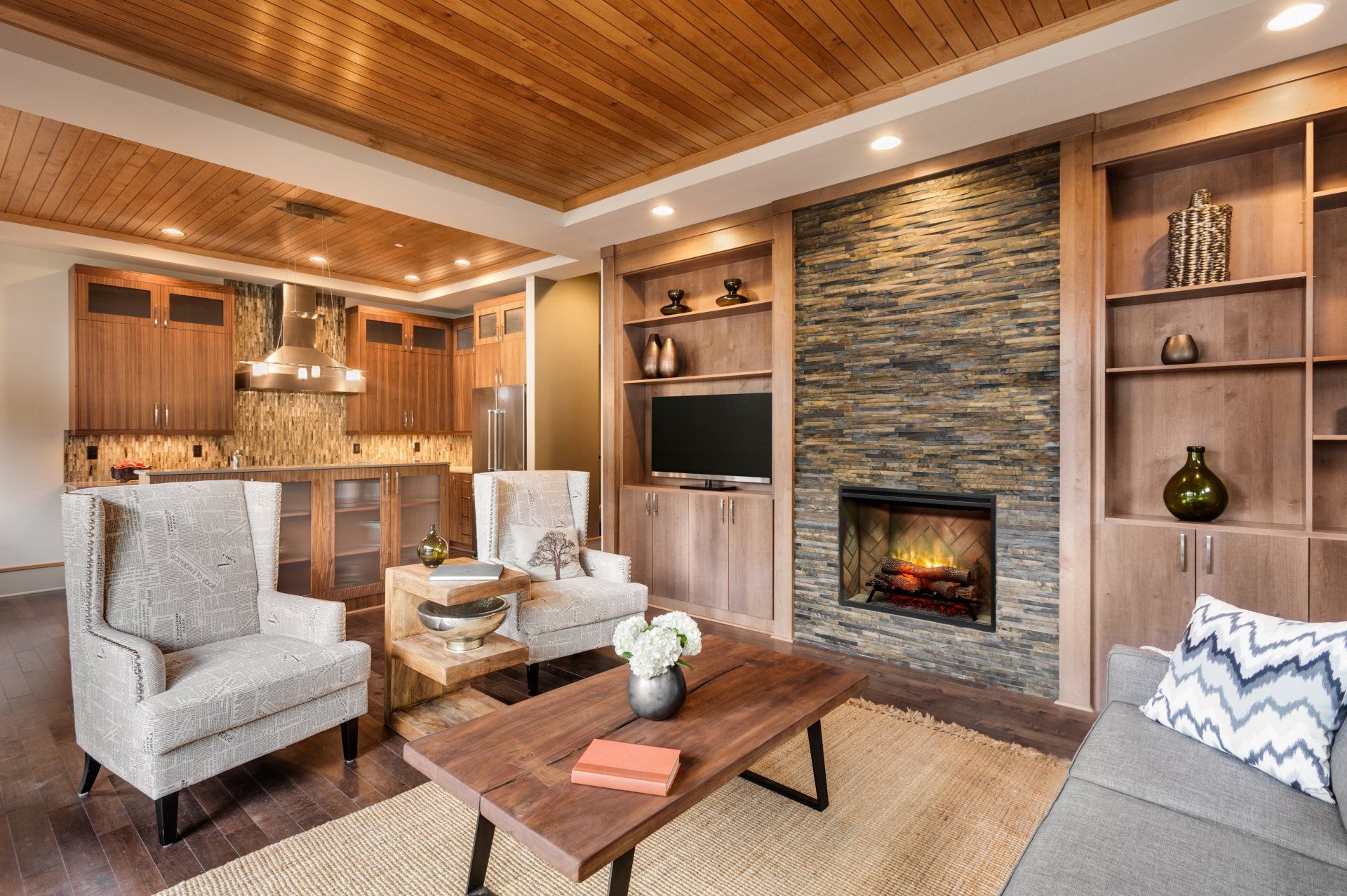 Cozy living room with fireplace, bookshelves, wood ceiling, chairs, and kitchen view.