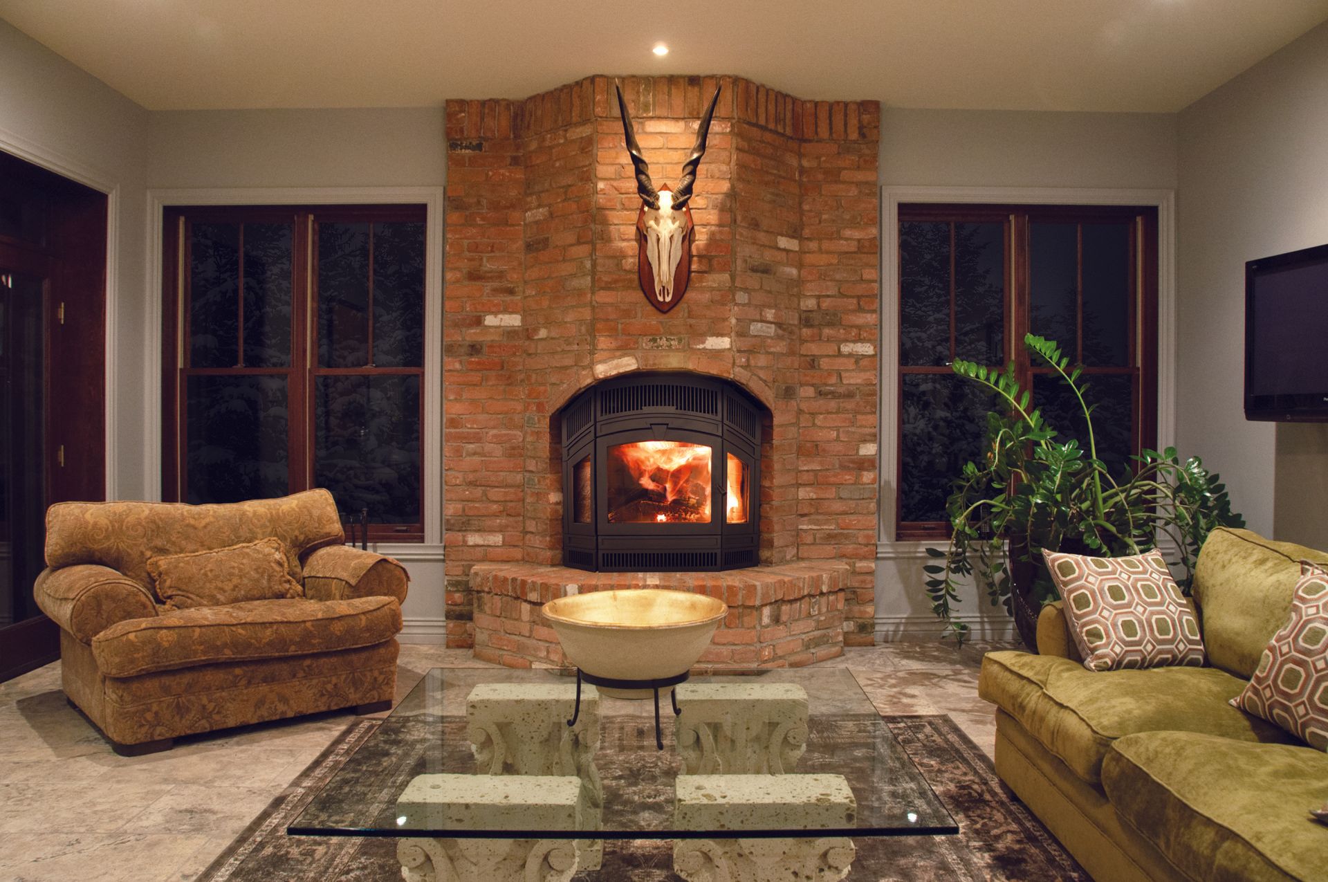Cozy living room with brick fireplace, antler mount, and plush furniture; natural light streams from windows.