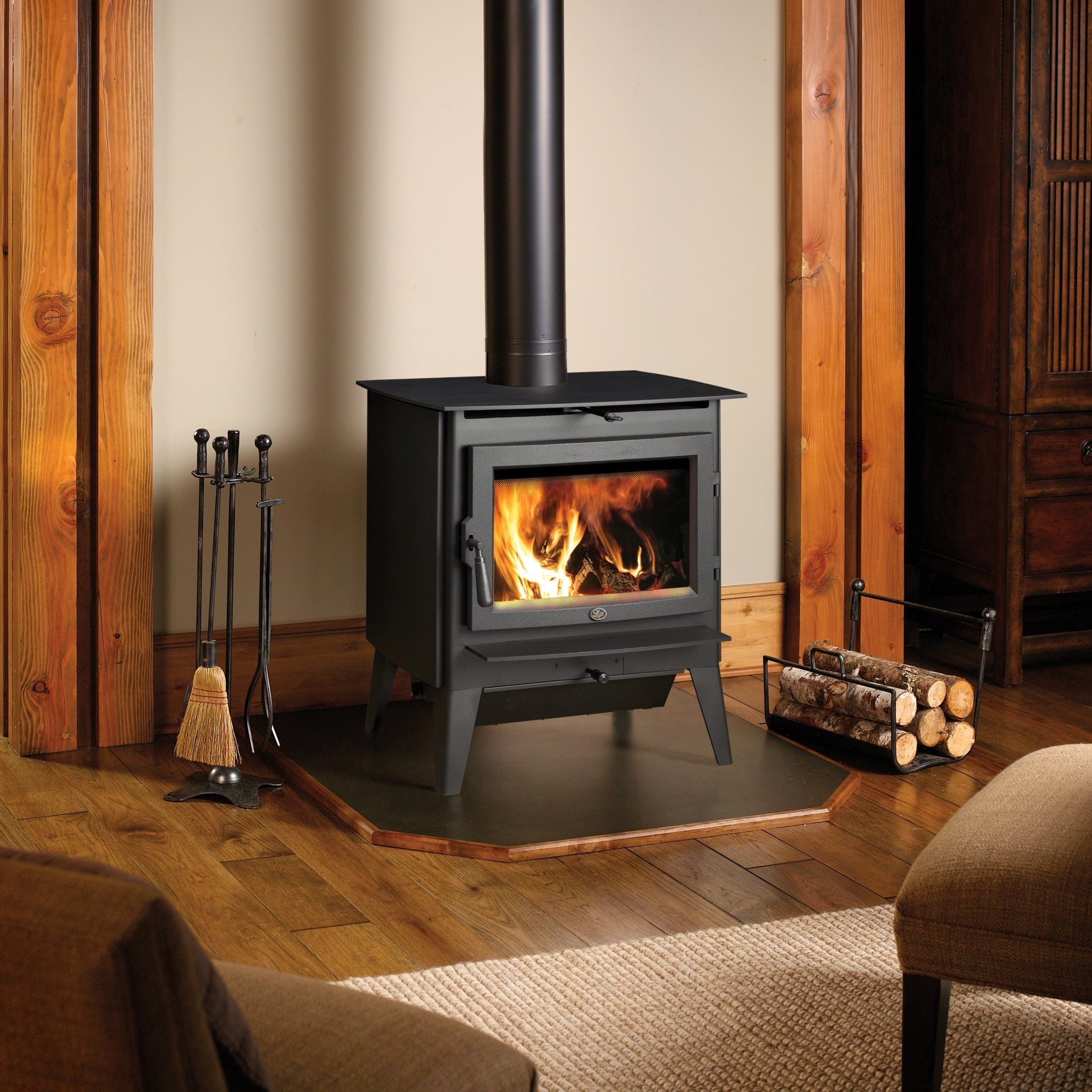 Black wood-burning stove with flames in a cozy living room setting. Fireplace tools and a wood rack are nearby.