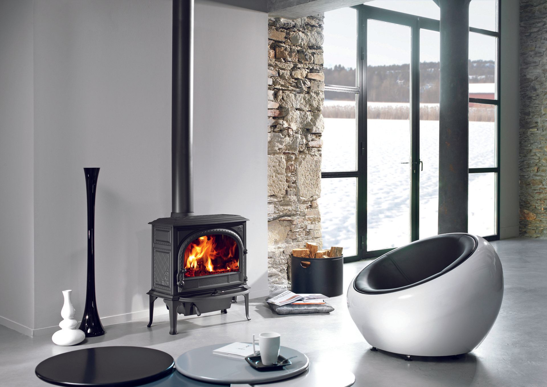 Cozy living room with a lit black stove, a modern white chair, and a view of water through a large window.