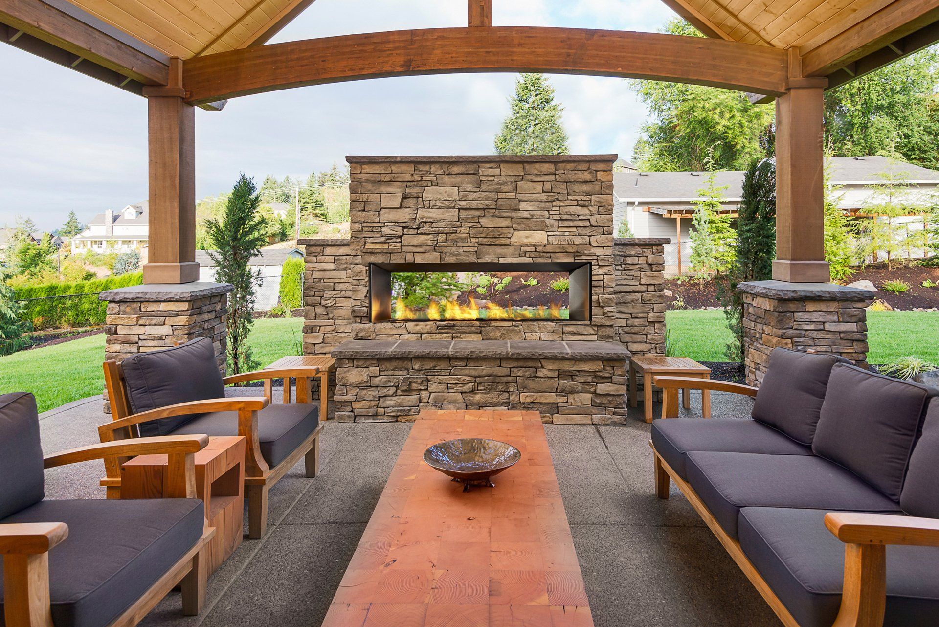 Patio with stone fireplace, wooden furniture, and outdoor seating under a covered structure.