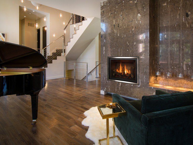 Living room with piano, fireplace, and staircase. Dark wood floors, dark green chair, modern decor.