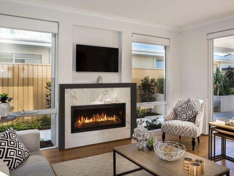 Living room with fireplace, TV above. Windows on either side, furniture, light wood floors, and neutral tones.