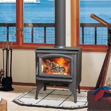 Wood-burning stove with flames in a room with a window overlooking water; hearth tools sit to the left.