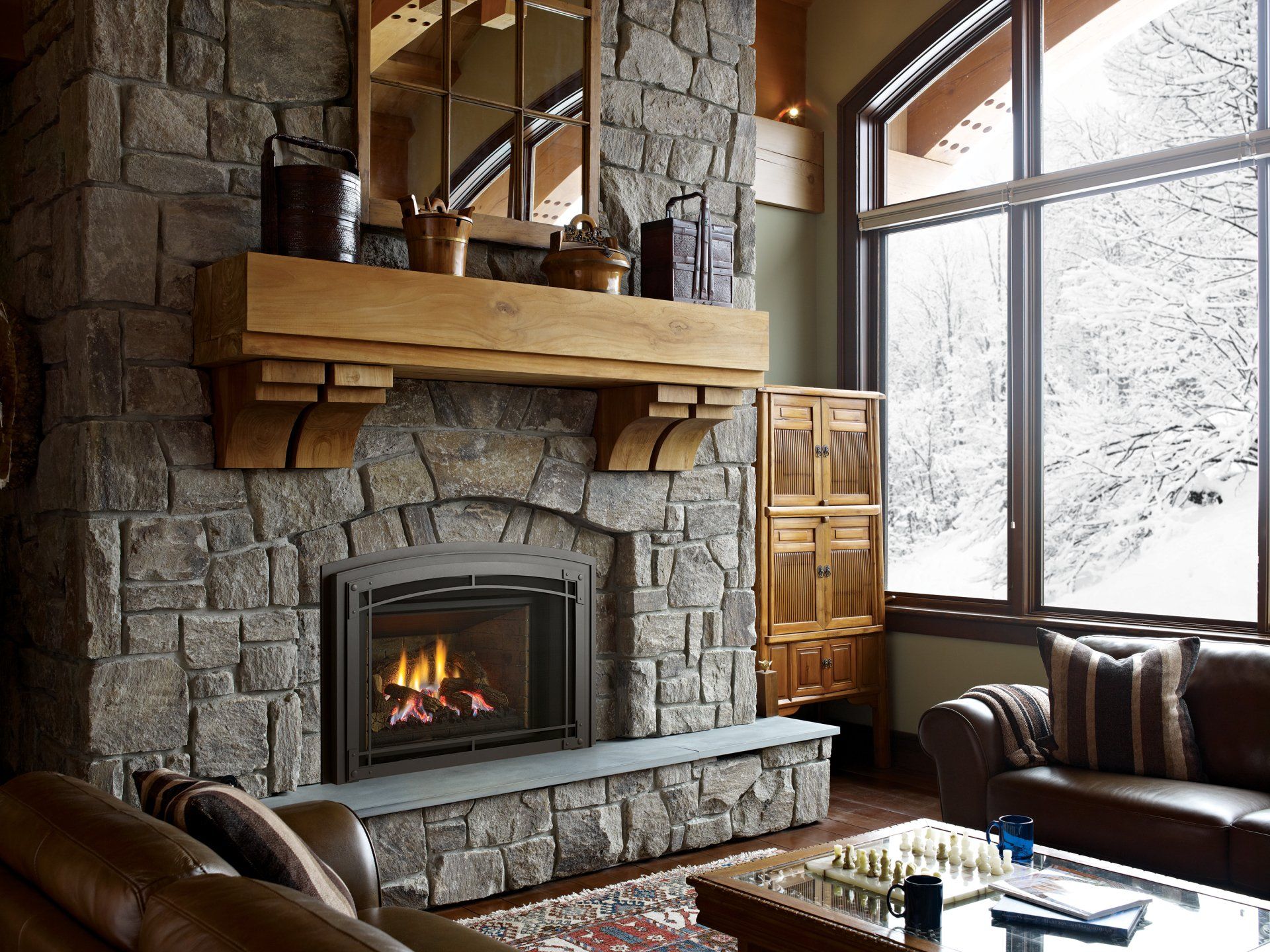 Cozy living room with stone fireplace, wood mantel, large window with snowy view, brown leather sofas.