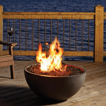 Fire pit on a wooden deck by water, flames burning, glass of wine nearby.