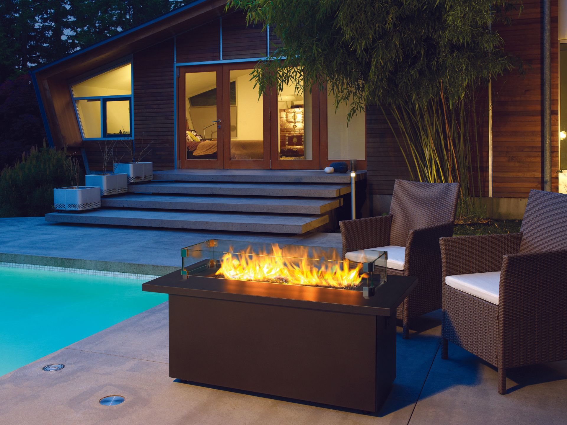 Fire pit with flames, wicker chairs, by a pool and modern house at dusk.