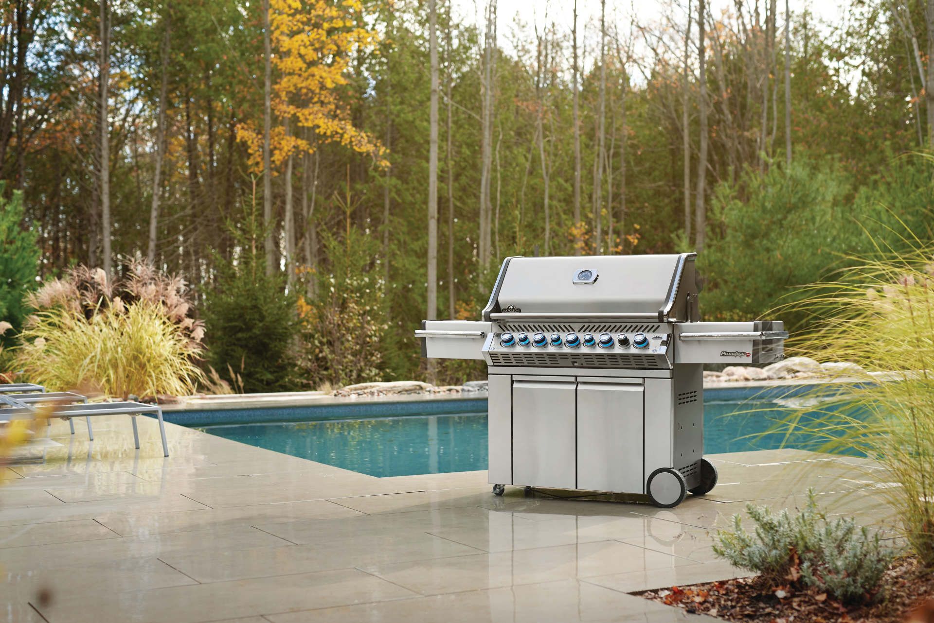 Stainless steel grill on a patio near a pool and trees.