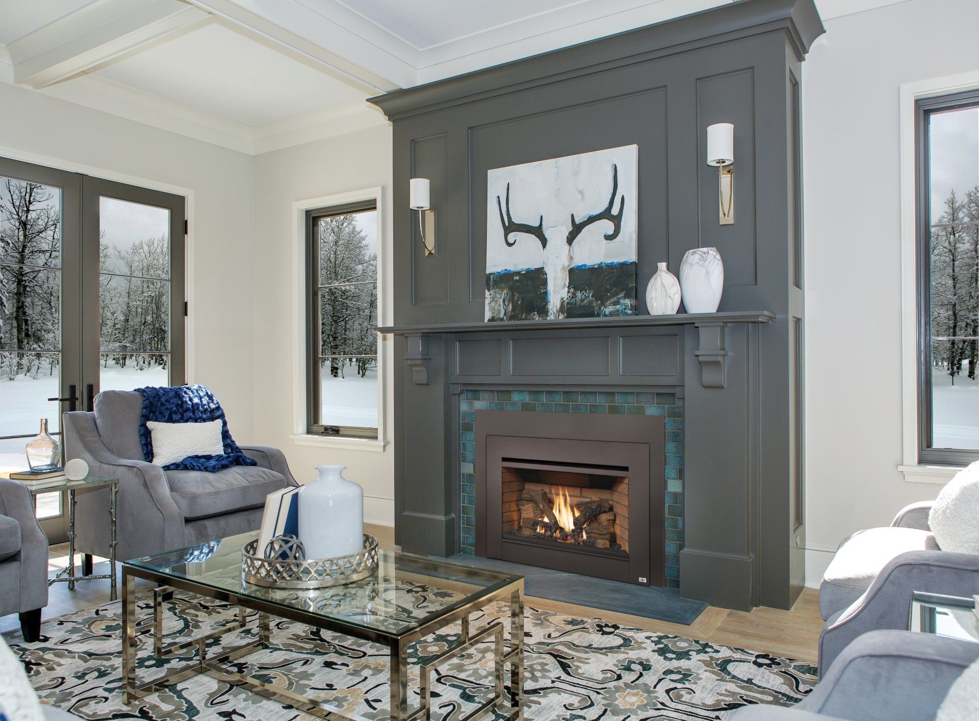 Cozy living room with a gray fireplace, chairs, and a glass coffee table, with snowy views outside the windows.