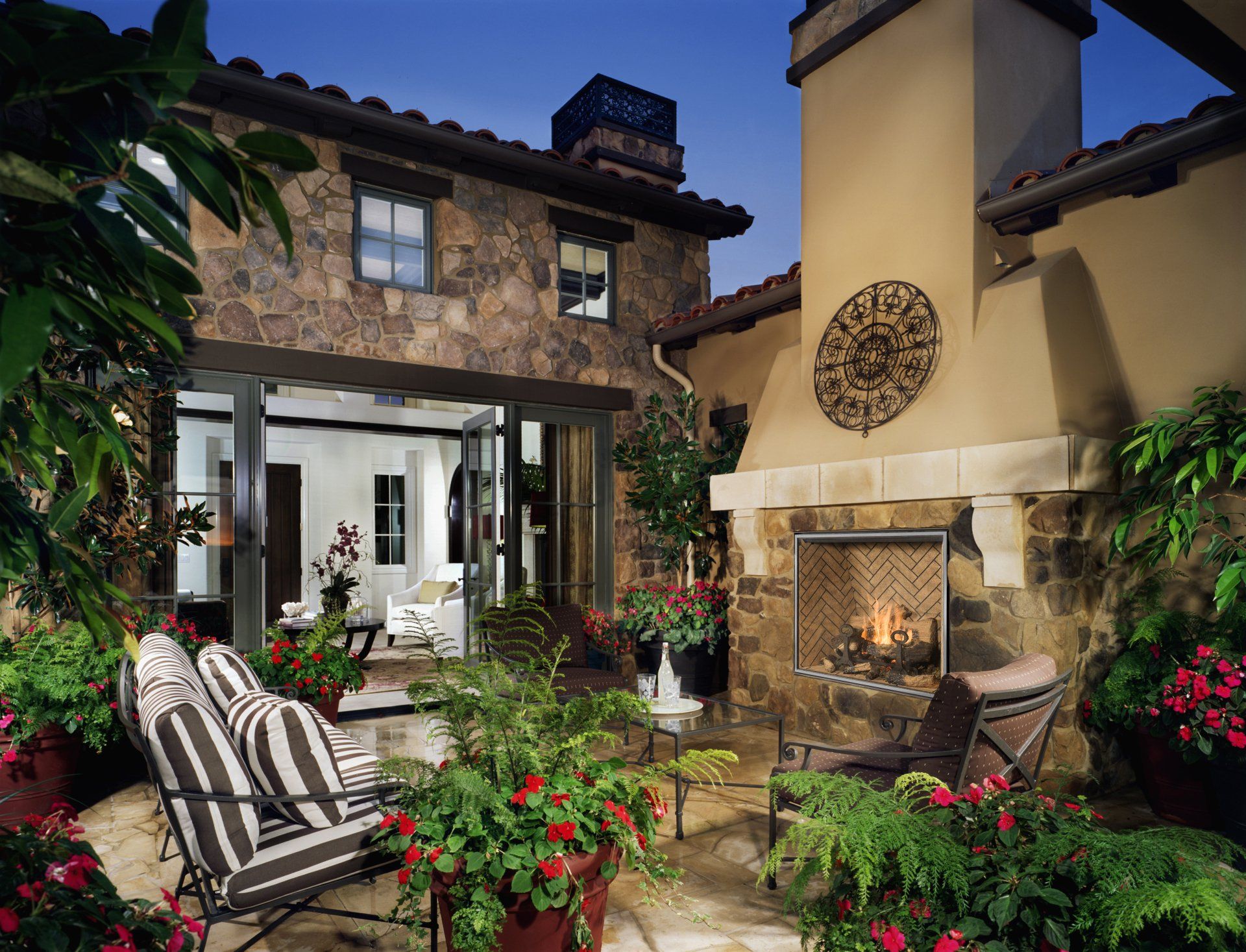 Stone patio with fireplace, seating, and lush greenery, leading to a home interior.