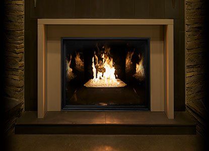 Fireplace with burning flames inside a rectangular glass enclosure, framed by a beige mantel, against a dark brick wall.