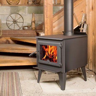 Black wood-burning stove with flames, chimney, in a wooden cabin setting.