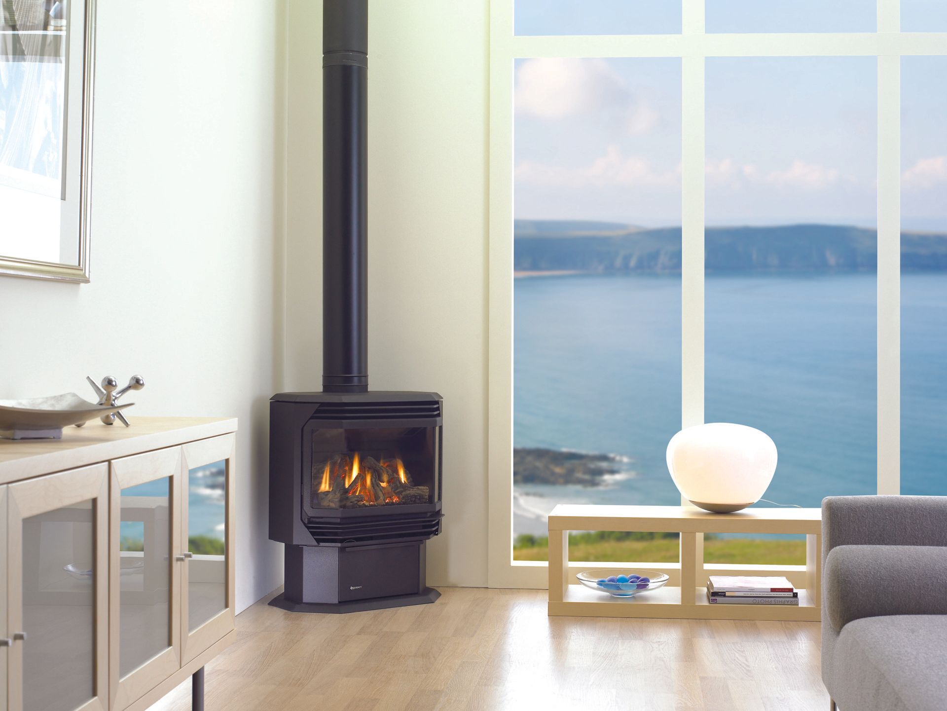 Black gas fireplace with tall chimney in a room with a large window overlooking the ocean.