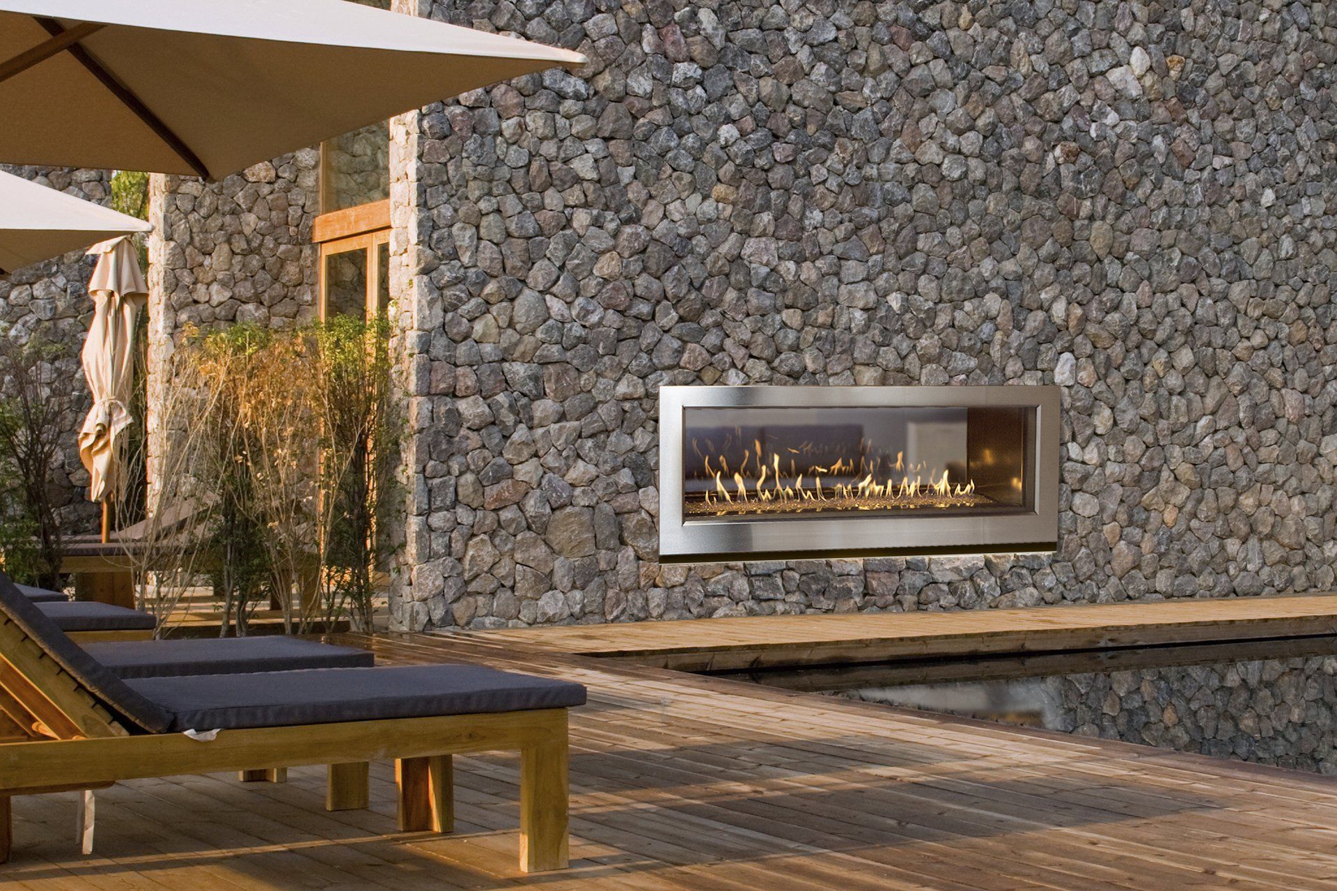 Outdoor fireplace built into a stone wall next to a pool and lounge chairs.
