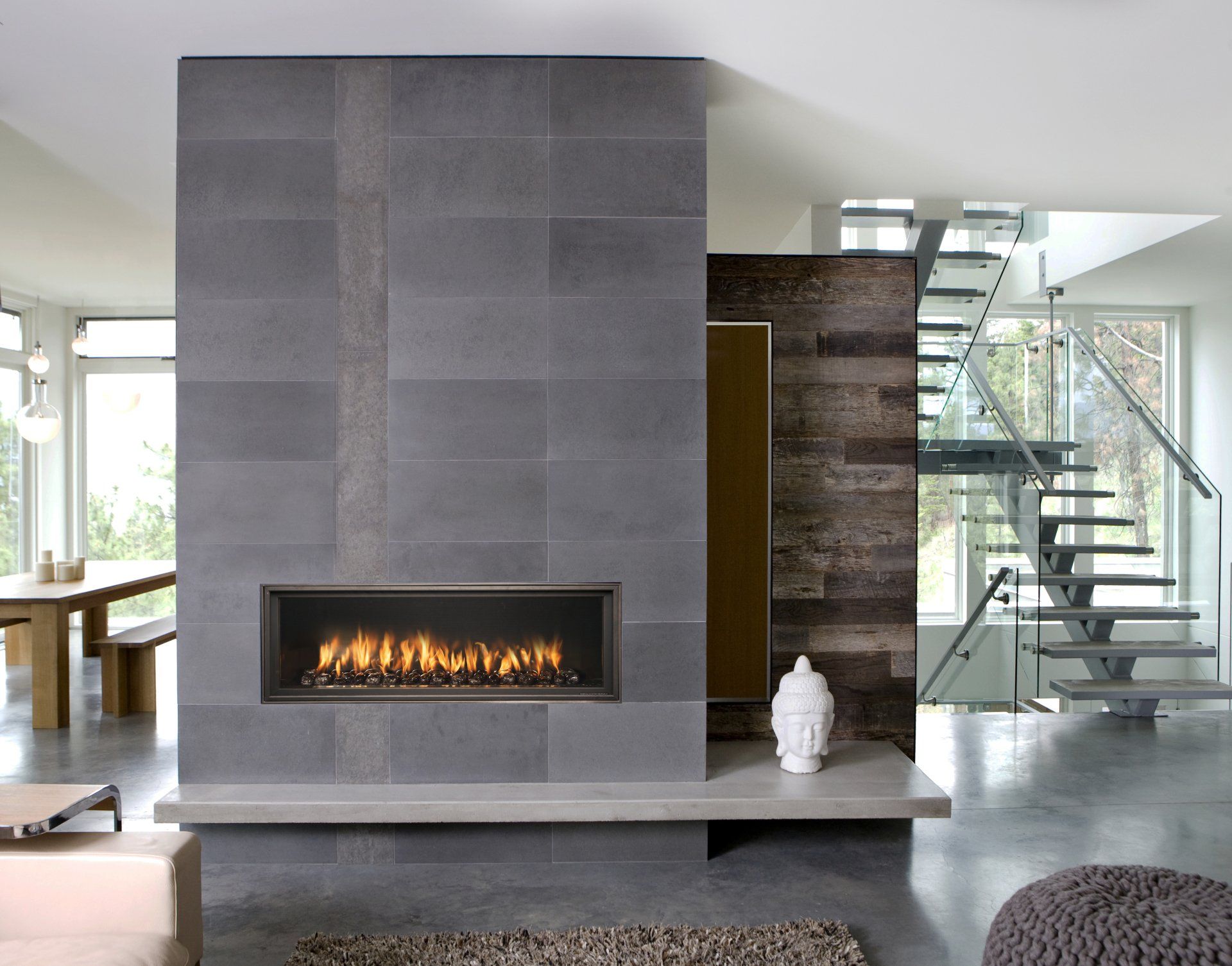 Modern living room with a gray tiled fireplace, flames, a floating staircase, and concrete floors.