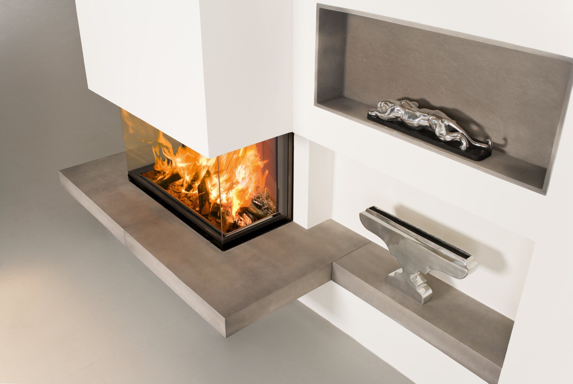 Modern fireplace with flames, built into a white wall. Shelves hold silver sculptures.